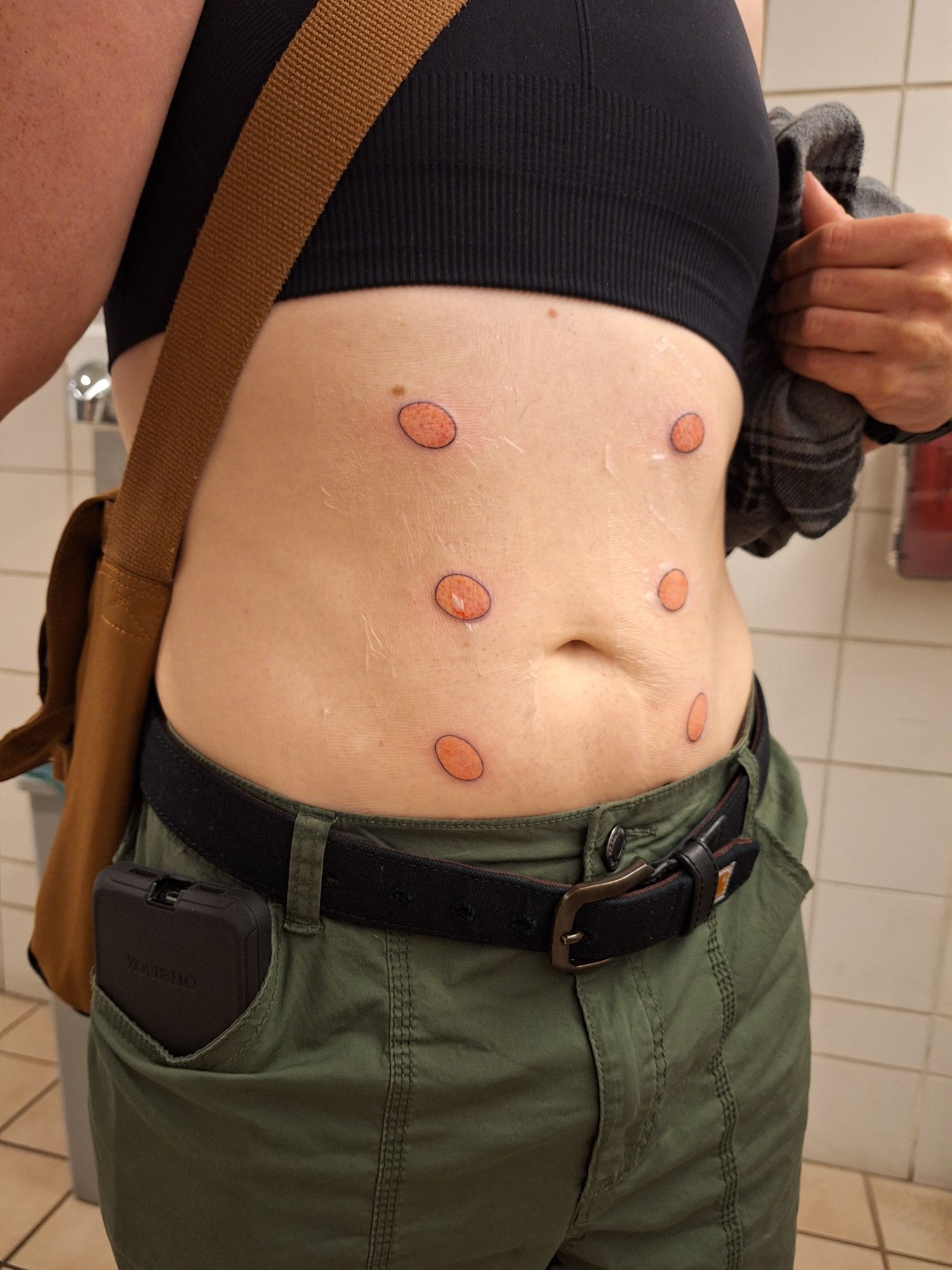 Juniper's torso taken in a convention center bathroom. She just got fresh tattoos of six abstract oval-shaped tummy areolas. They have a thin black outline with a pink interior, and follow an hourglass shape from the bottom of her ribcage to just above her waistband.