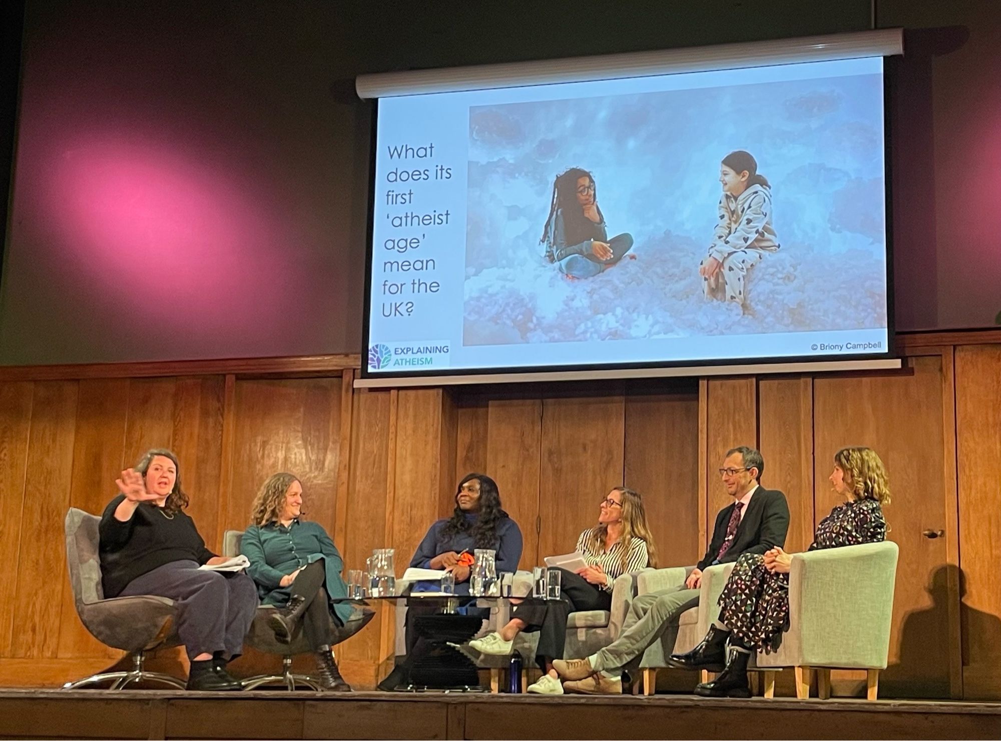 Panel of 6 people sitting on a stage with PowerPoint slide behind featuring two children having a discussion while apparently sitting on clouds