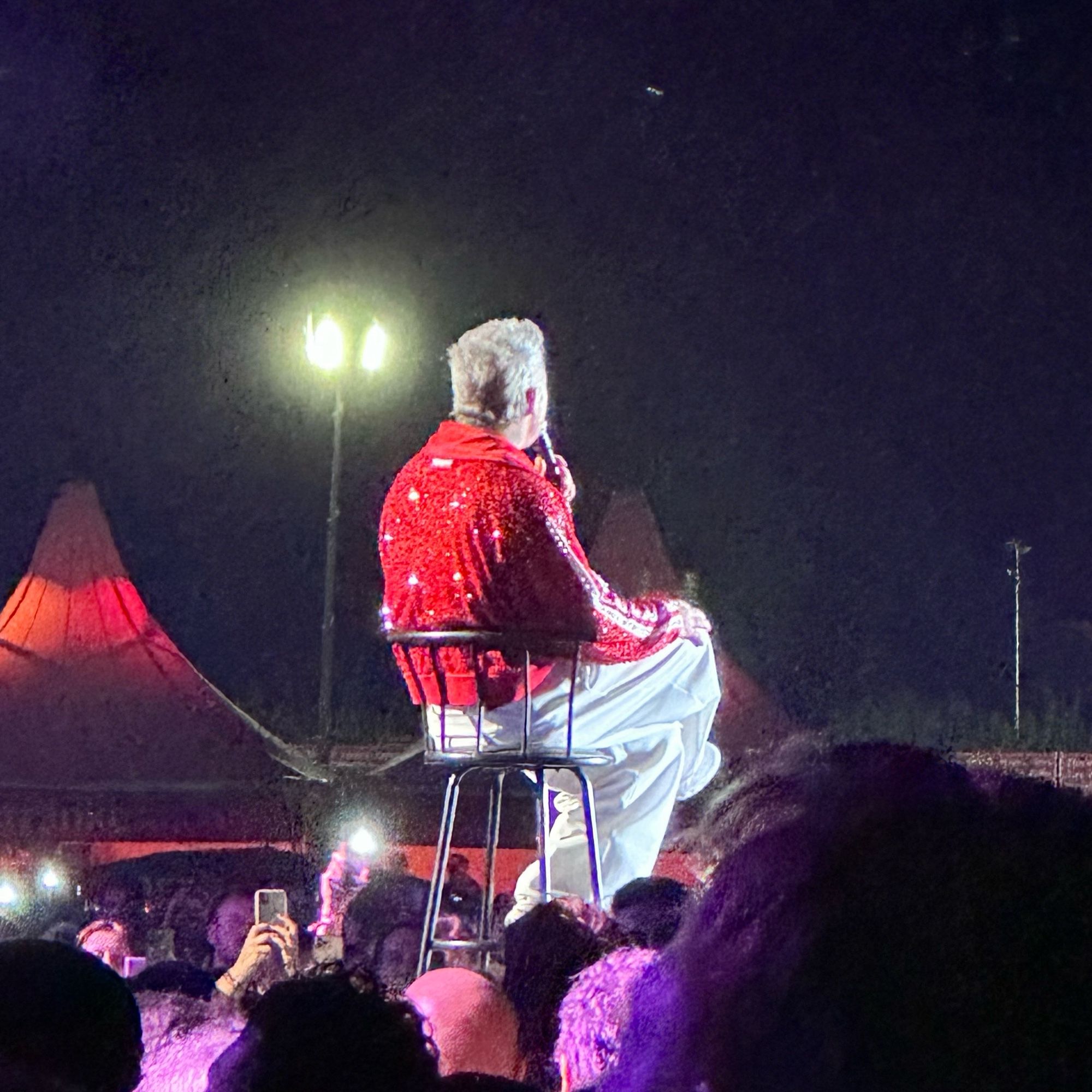 Robbie Williams sitzt mit roter Glitzerjacke auf einem Stuhl beim Konzert auf der Trabrennbahn in Hamburg.