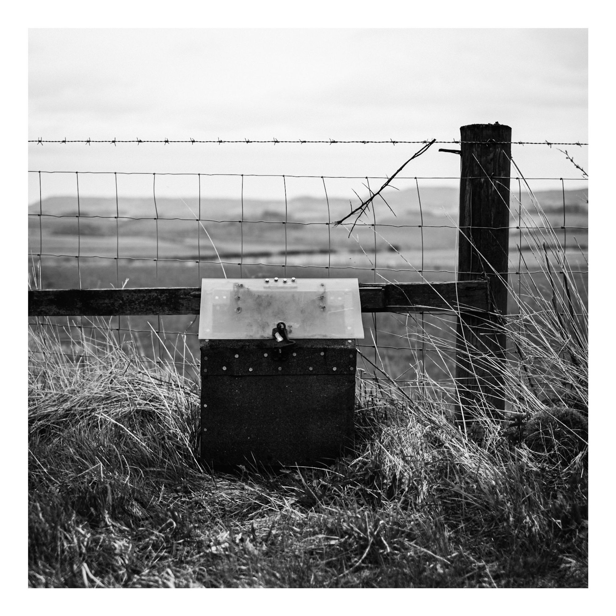 A black and white photograph featuring a small locked newspaper drop box placed near a barbed wire fence, with grassy terrain and distant hills in the background.