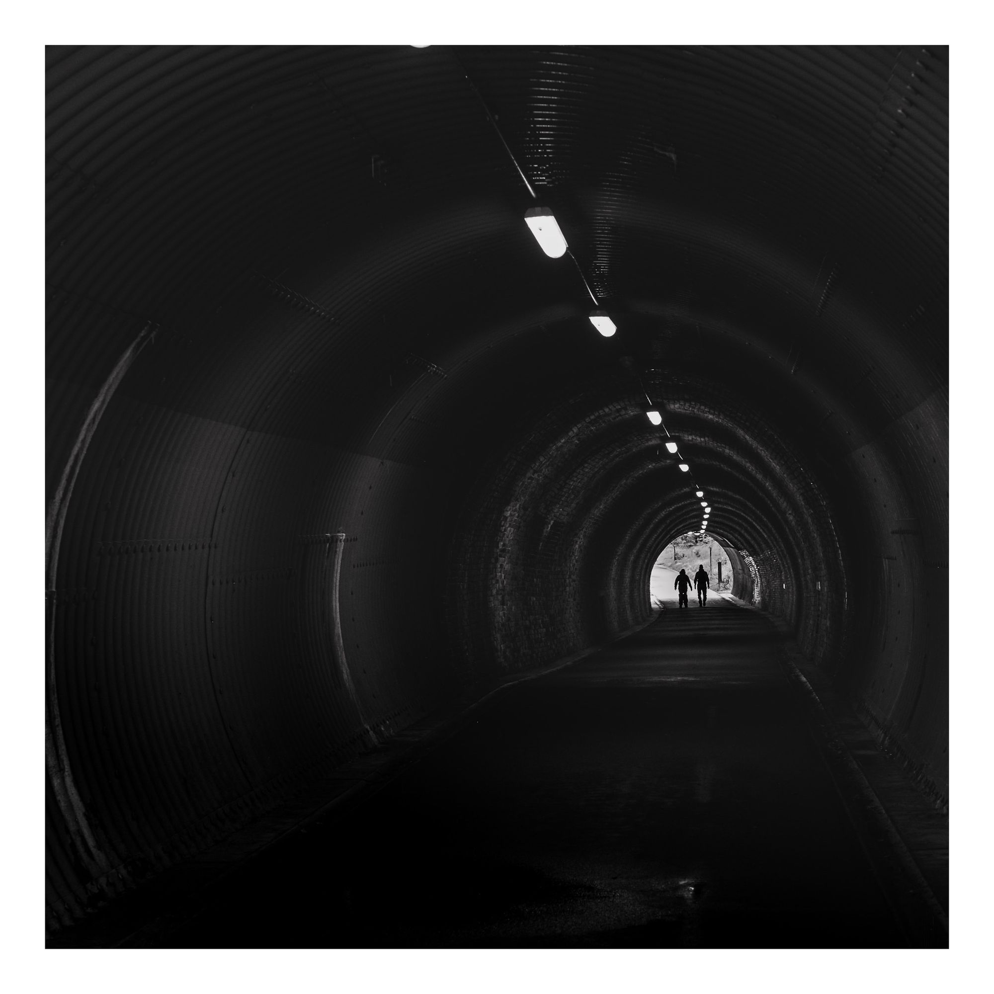 A dimly lit tunnel with a smooth, curved interior. Two silhouetted figures are walking towards the light at the end of the tunnel. The scene is monochromatic, emphasizing the contrast between light and dark.