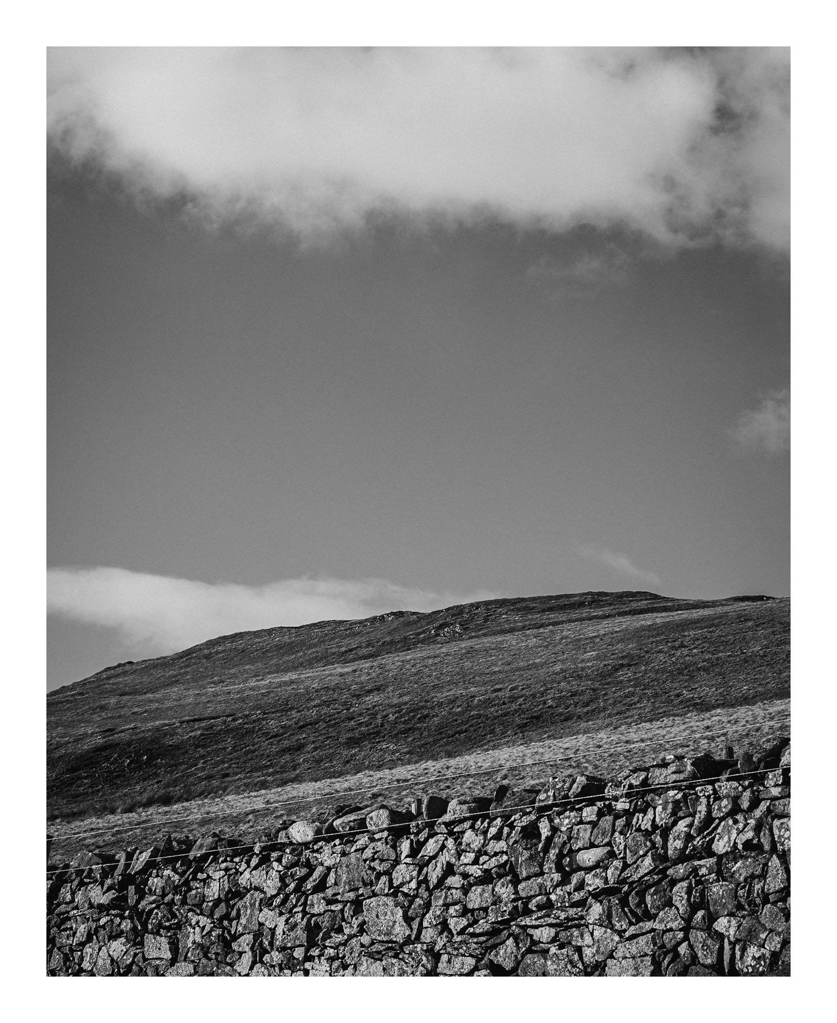 A black and white landscape featuring a stone wall in the foreground and a grassy hill beneath a cloudy sky in the background.