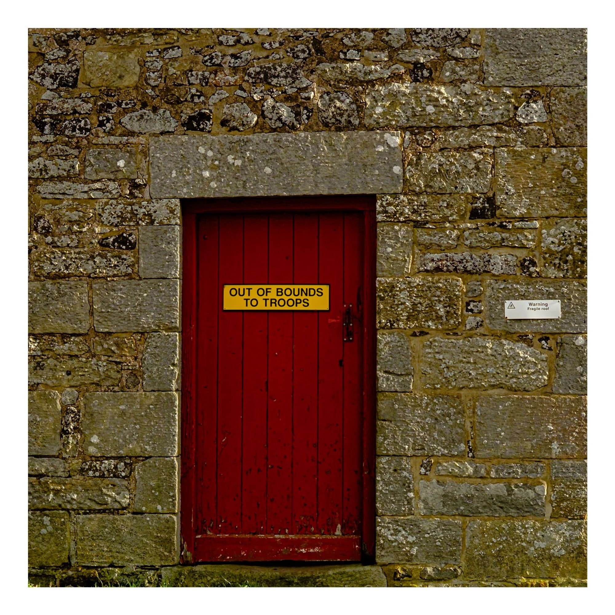 A red exterior door with a sign reading "OUT OF BOUNDS TO TROOPS," set against a stone wall. A smaller warning sign is “WARNING FRAGILE ROOF” is visible to the right of the door.