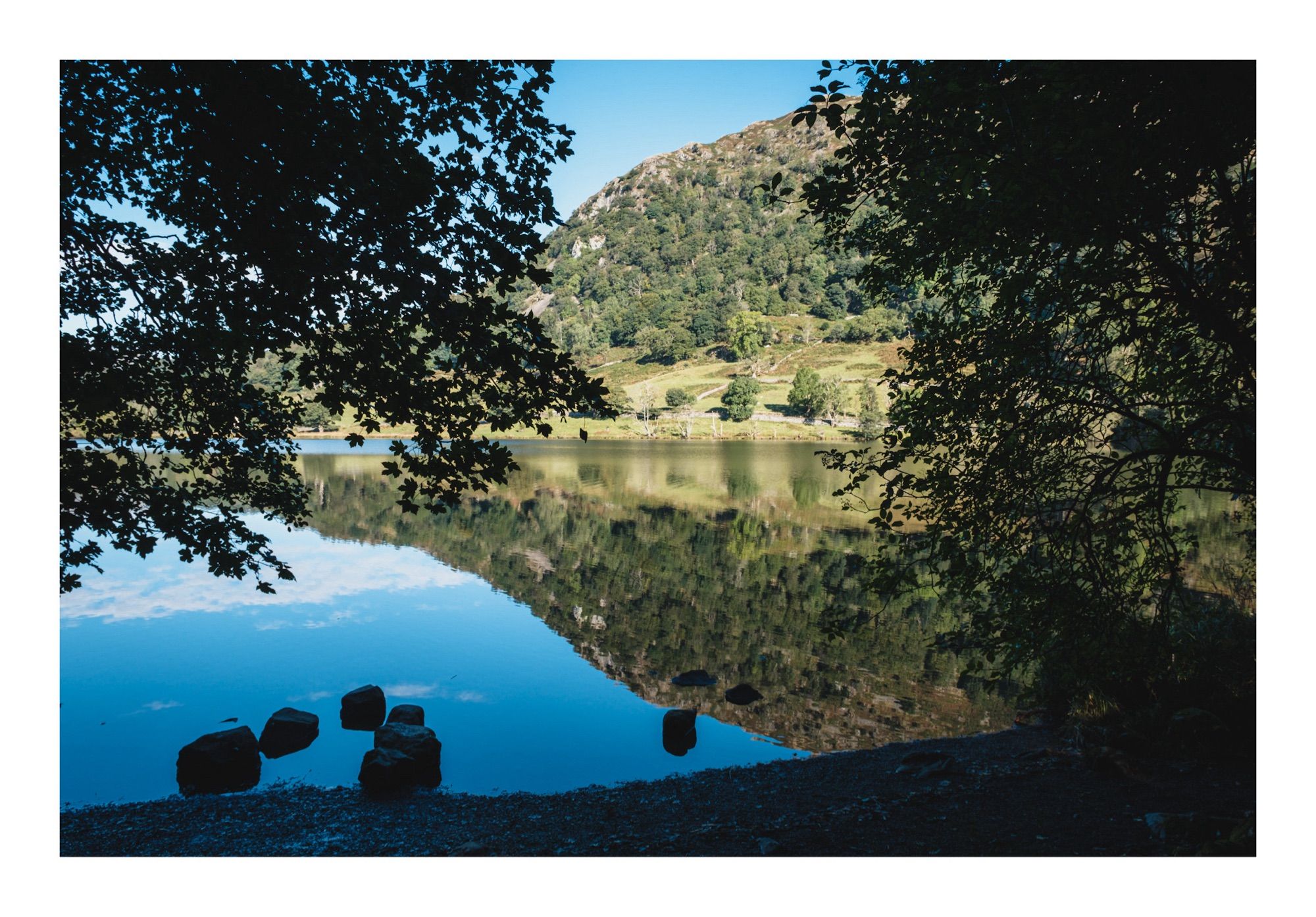 Trees overhang a lakeside with a rocky hillside reflected in the lake.