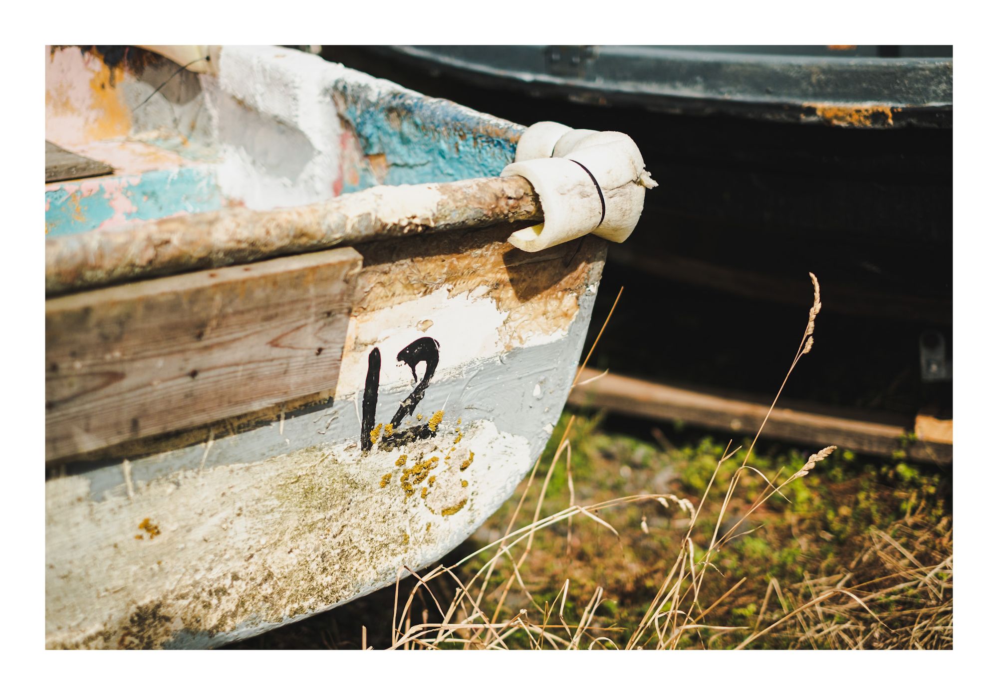 A colour photograph of the close up of a boat keel with the number 12 on it in black paint. The wood is aged and bleached he’s and some grass stalks are in the foreground. 