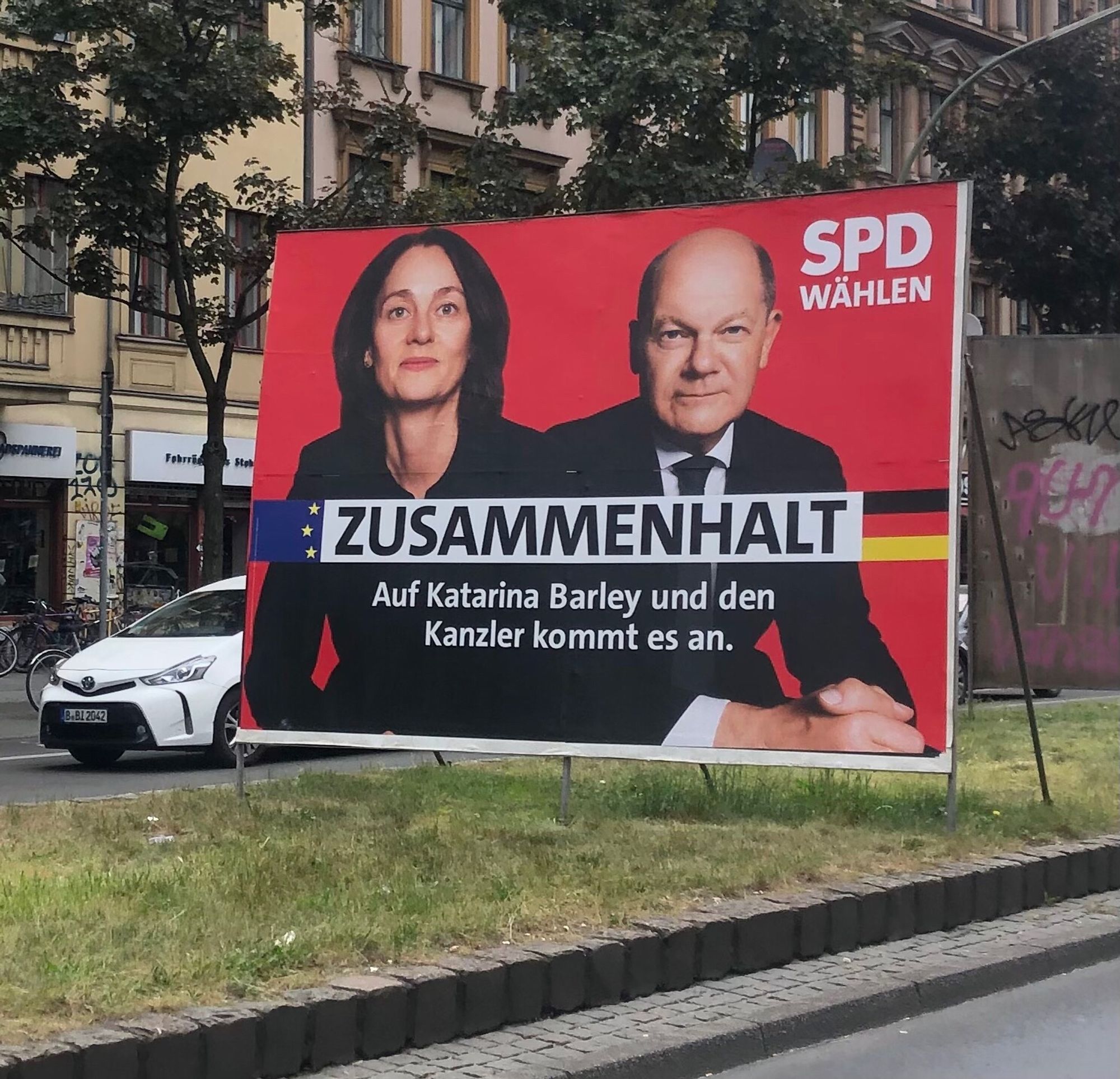 Foto eines Wahlplakates der SPD. Darauf sind links Katharina Barley und rechts daneben Olaf Scholz abgebildet. Der Slogan: ZUSAMMENHALT
Auf Katarina Barley und den Kanzler kommt es an. 
Das Plakat ist auf einer Plakatwand und diese steht auf einem Grünstreiifen zwischen zwei Straßen.