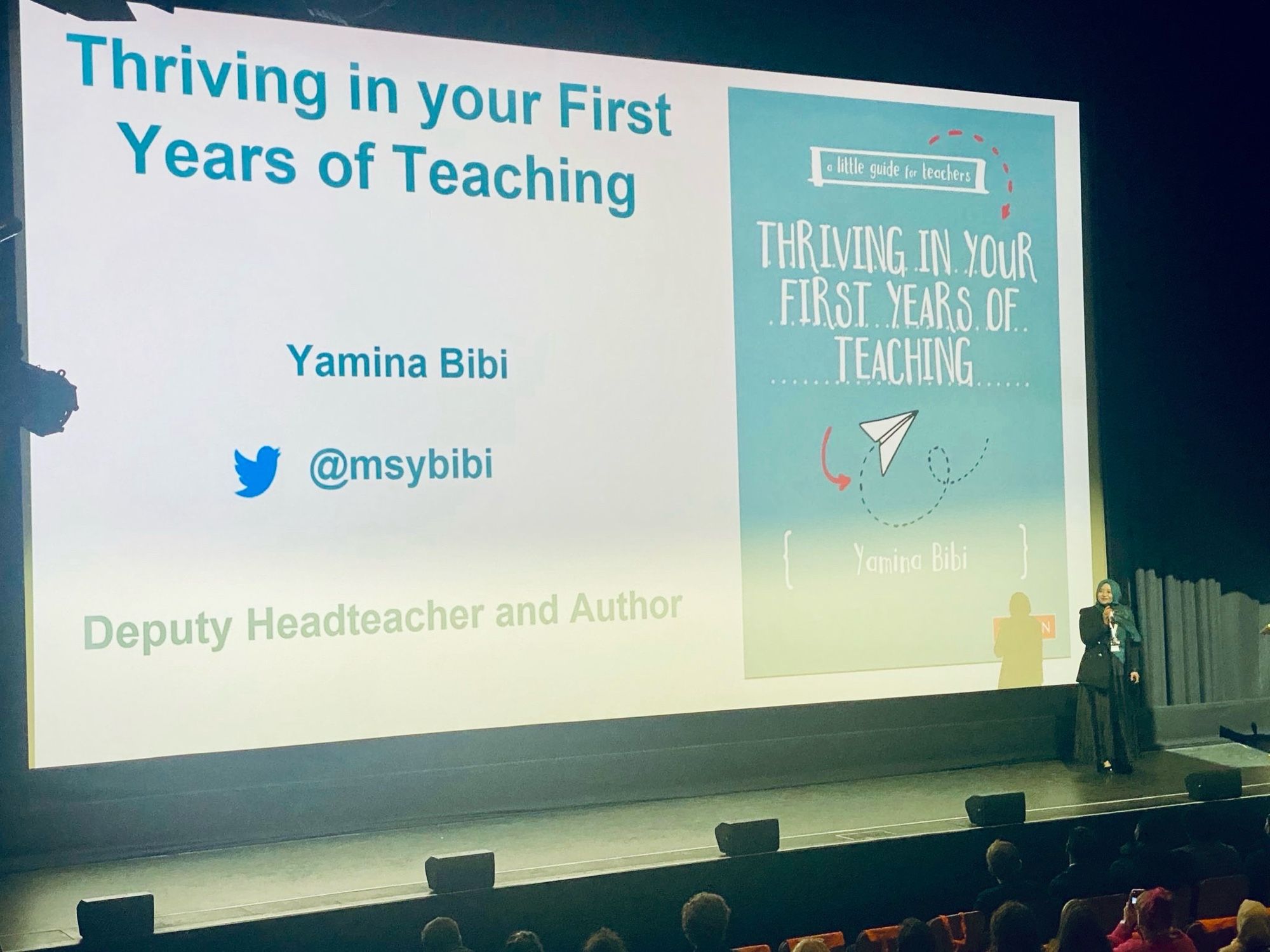 Image of Yamina standing a stage. 
There is a large screen behind her with her book cover and title reads ‘Thriving in Your First Years of Teaching’