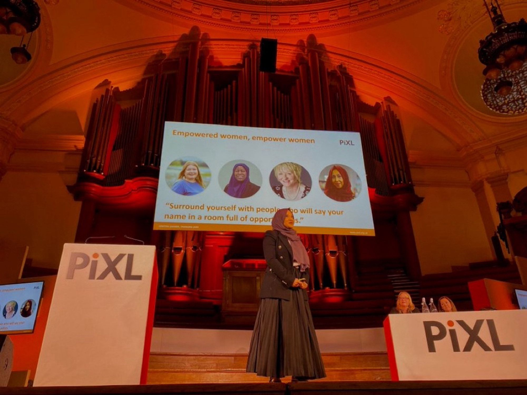 Yamina standing on a stage wearing a black blazer, scarf and black skirt. There is a lectern which has PiXL written on it. 
Behind her is a screen with pictures of 4 women.