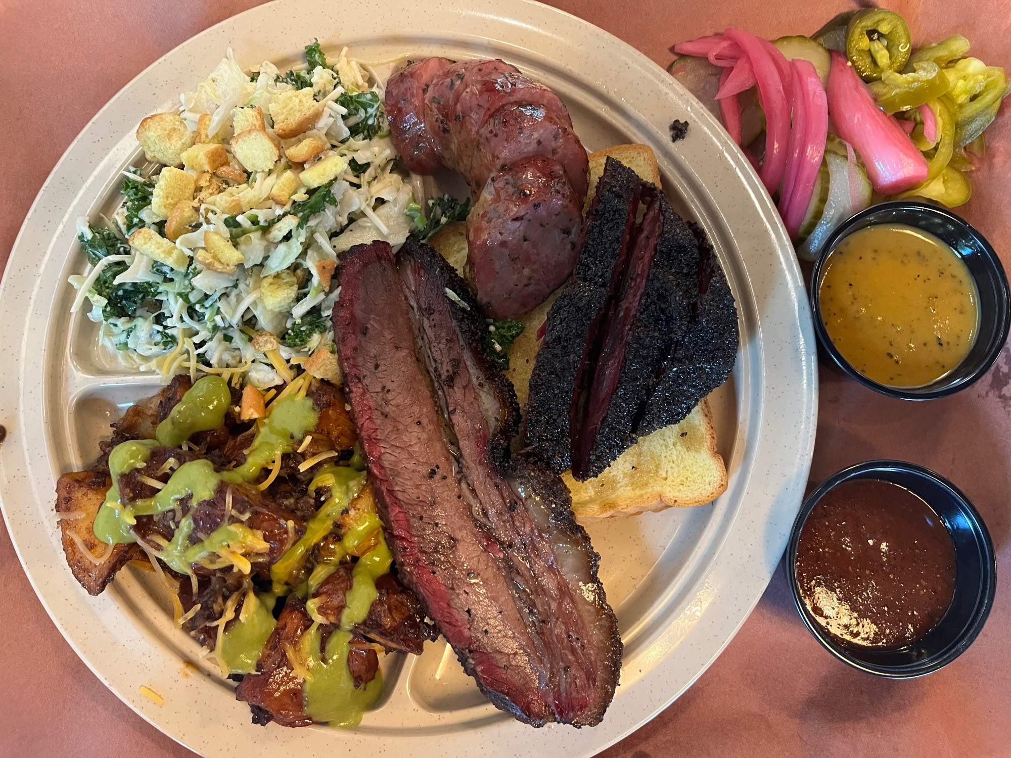 To the left of the photos is a plate of “new school” barbecued meats at LeRoy & Lewis in Austin, Texas: a slice of brisket, sliced pork-and-hop sausage and sliced beef cheeks covered with a dark “bark” coating, with two sides: kale caesar slaw and chori-papas (chorizo potatoes) topped with green salsa. Two small tubs of barbecue sauce and loose pile of pickled onions and jalapenos sit on the right side of the tray.