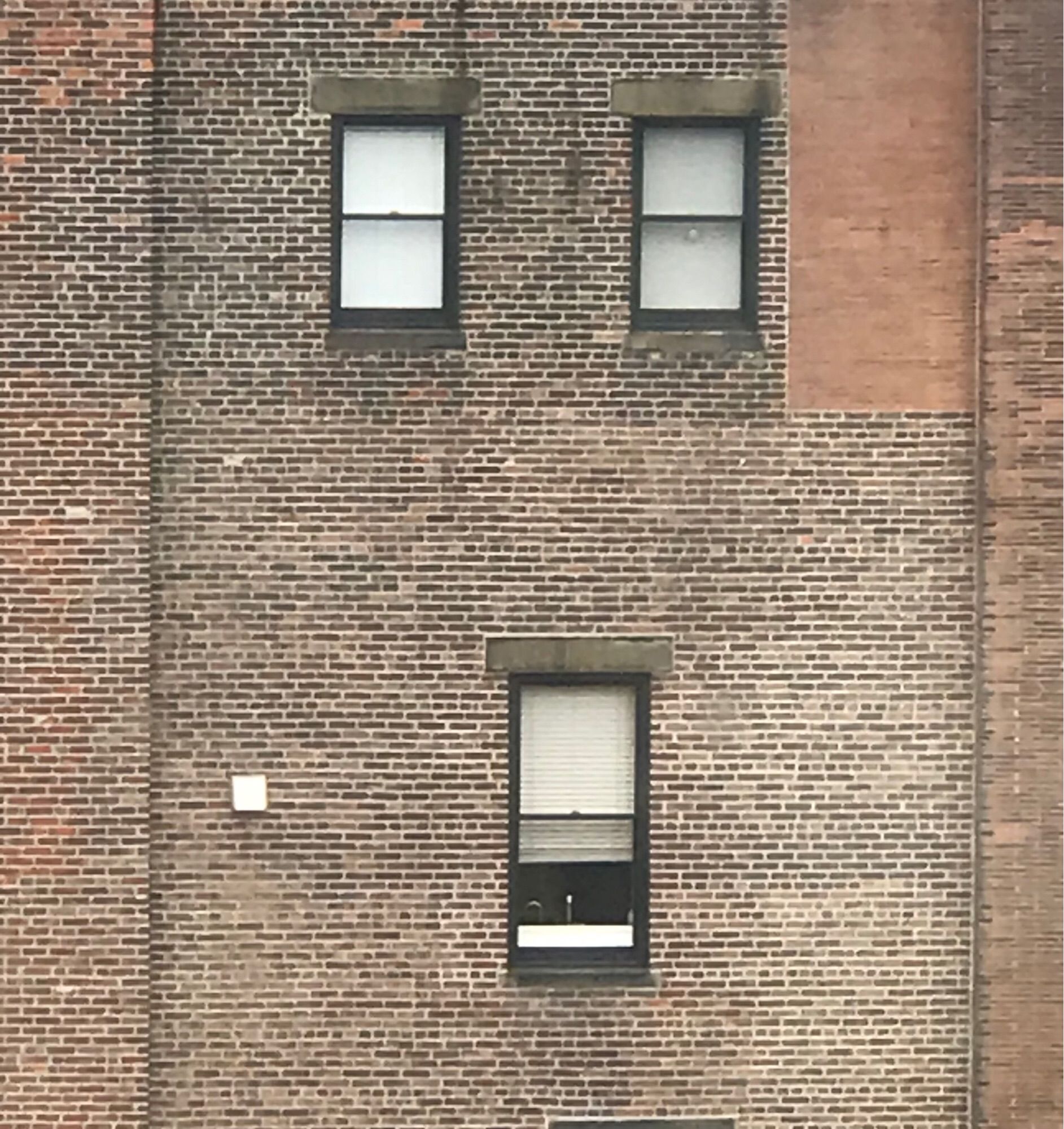 Side of a brick building. Three windows, two in a row and one beneath and between them, with shades drawn. The window on the bottom’s shade is 2/3 drawn.