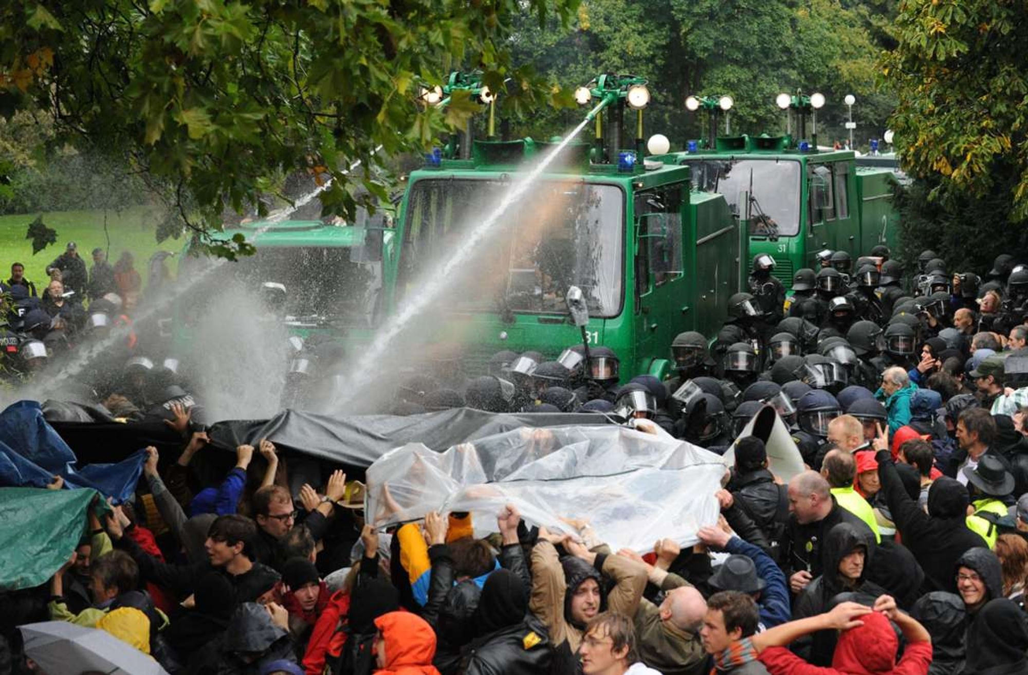 Mehrere Wasserwerfer hintereinander umringt von der Polizei bahnen sich einen Weg durch dicht an dicht stehende Menschen die sich zum Teil versuchen mit Planen zu schützen.