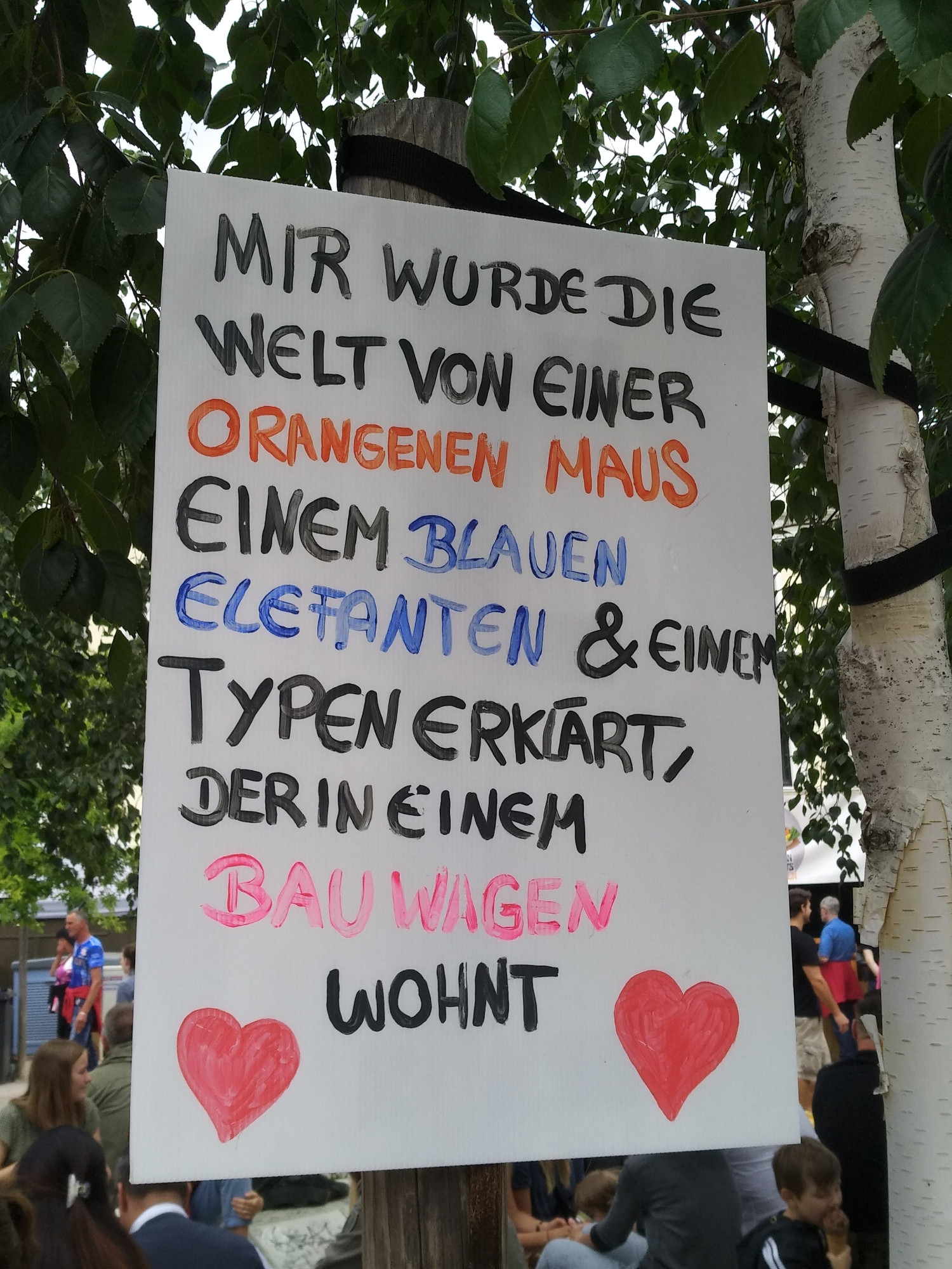 A0 großes, weiße Pappe ist an den Stützpfosten eines Baumes gehängt. Darauf mit Edding geschrieben: Mir wurde die Welt von einer orangenen Maus einem blauen Elefanten & einem Typ erklärt, der in einem Bauwagen wohnt.