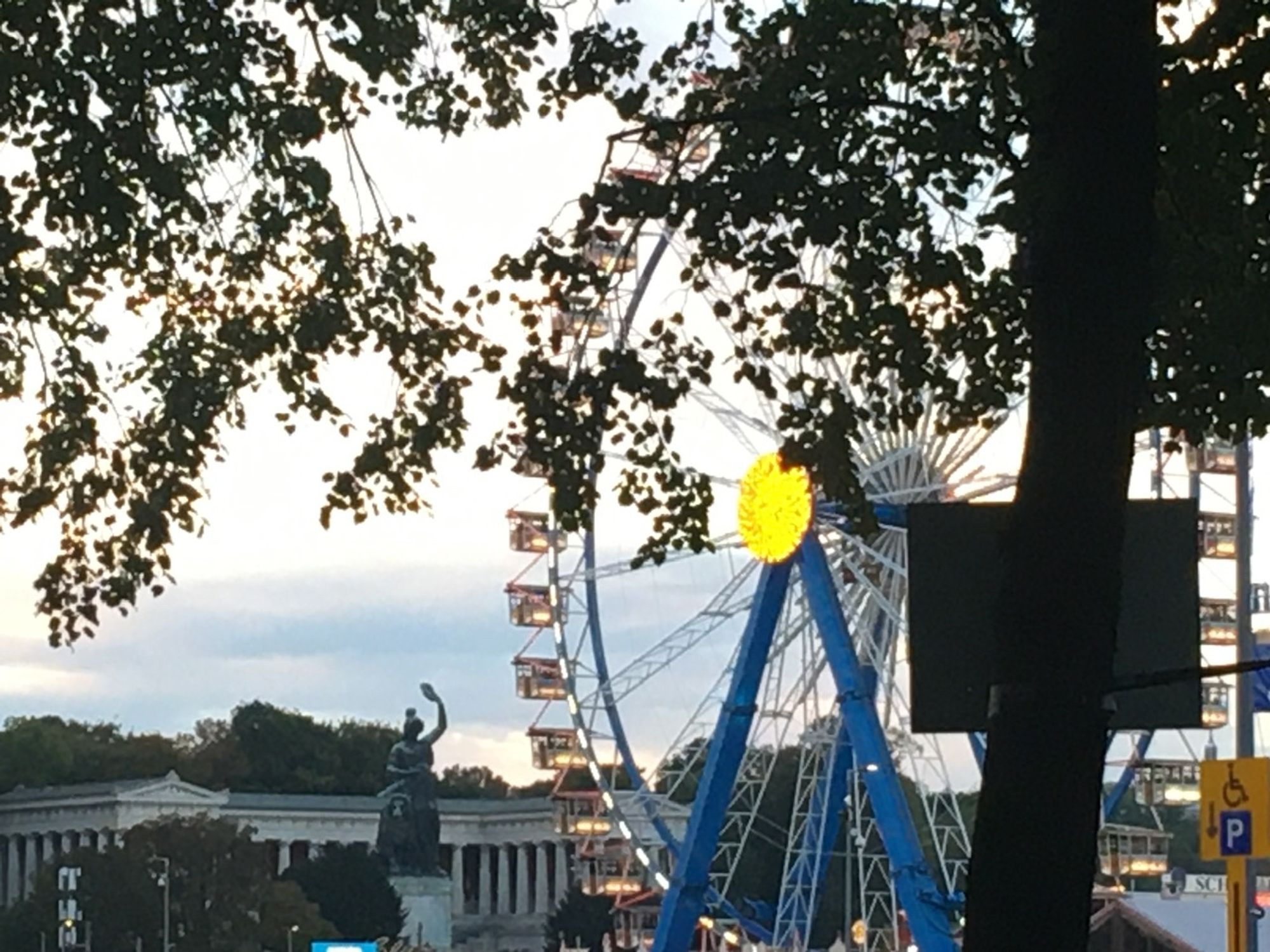 Im Vordergrund das Riesenrad gelb leuchtend in der Mitte, im Hintergrund die Bavaria vor der Ruhmeshalle im klassizistischen Stil.