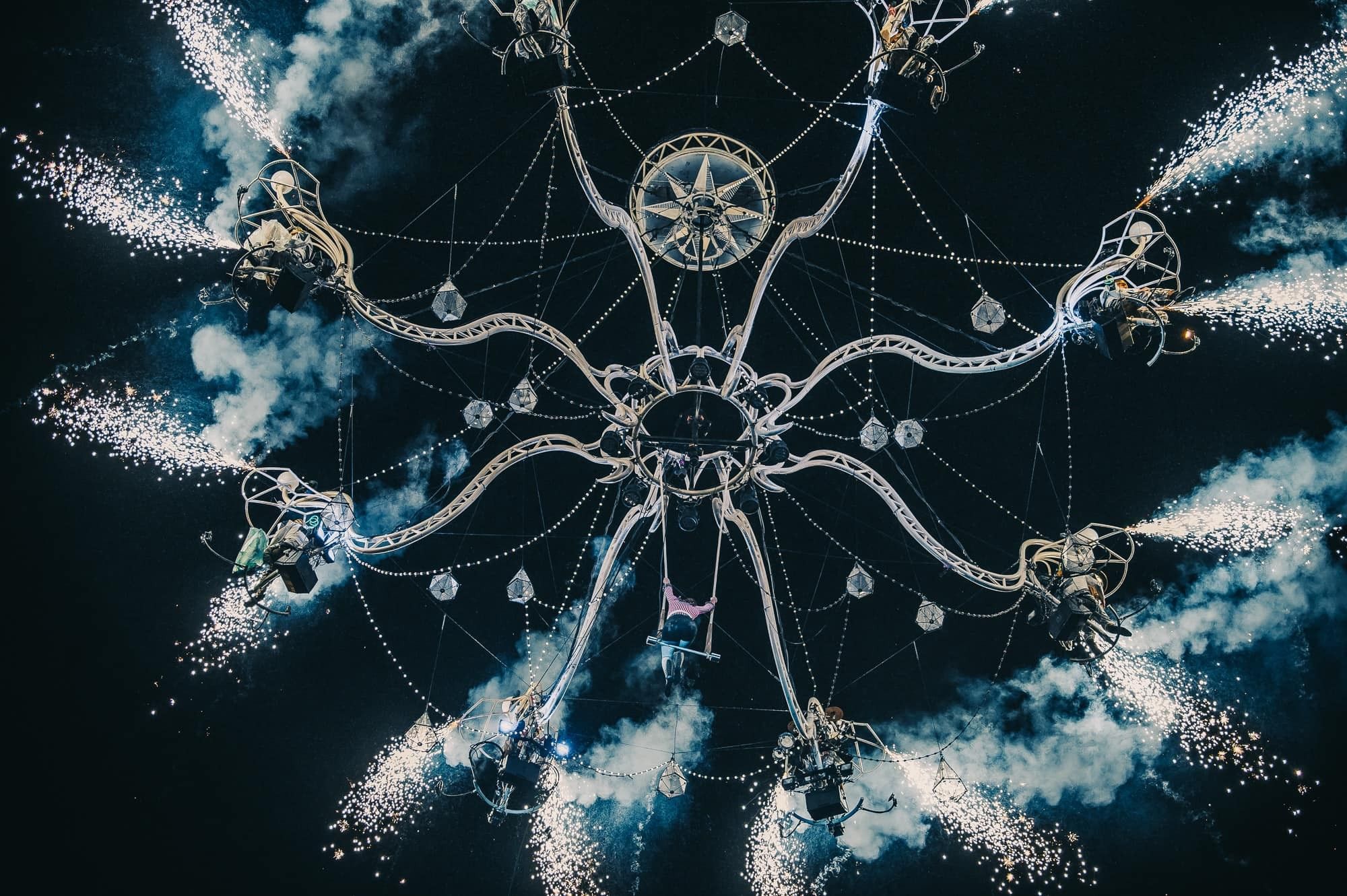 Photo d'un lustre géant avec des pampilles accroché pour un dispositif de spectacle 
A l'extrémité de chaque branche de lustre une place est prévue pour un artiste, au centre du lustre un trapèze
La photo est prise de nuit et les artistes laissent échapper des fumigènes blancs donnant l'impression de poussières d'étoiles