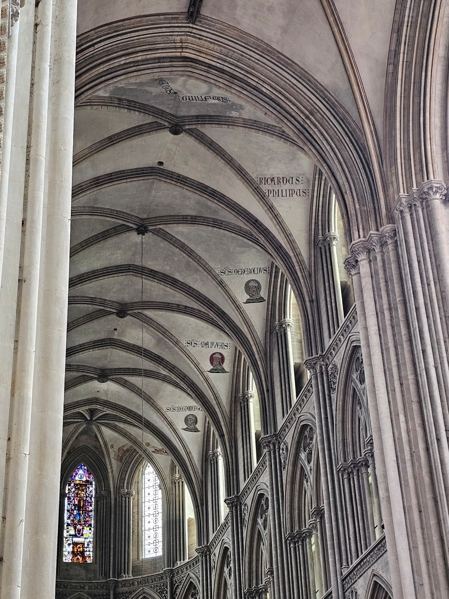 The tall vaulted archways of the inside of a gothic cathedral, leading to a stained glass window at the far end. Along some of the arches are portraits of different saints