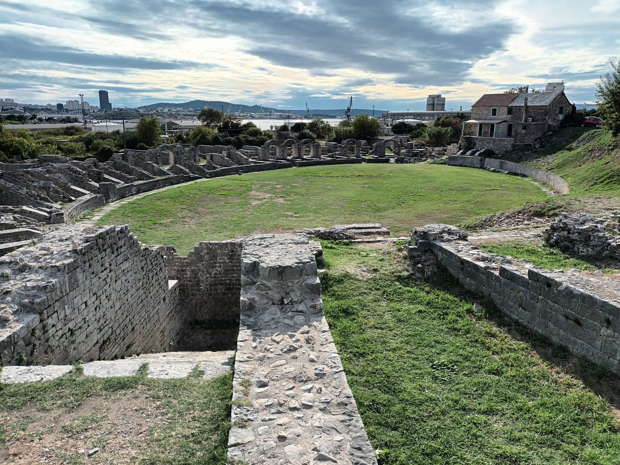 The circular ruins of a Roman amphitheatre with green grass in the centre, with a modern house on the right and modern city in the distance