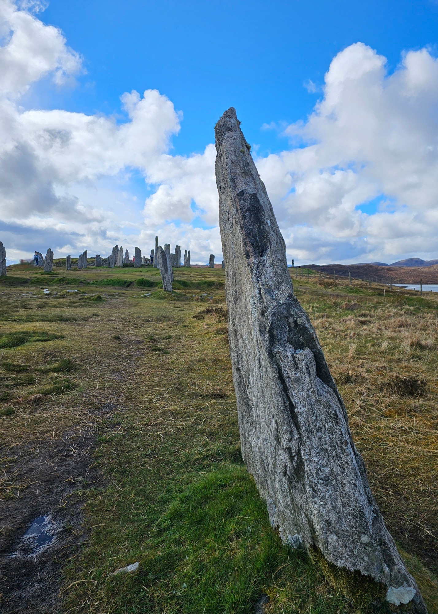 A grey angular standing stone embedded in mossy and grassy earth next to a track leading up to a cluster of standing stones in the distance, under a blue and white cloudy sky 