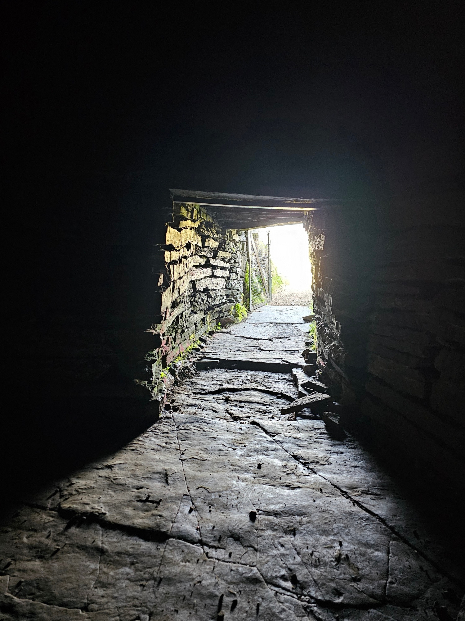 A dark image taken inside a stone tomb lookkng outwards down a narrow stone-lined passageway to the exit