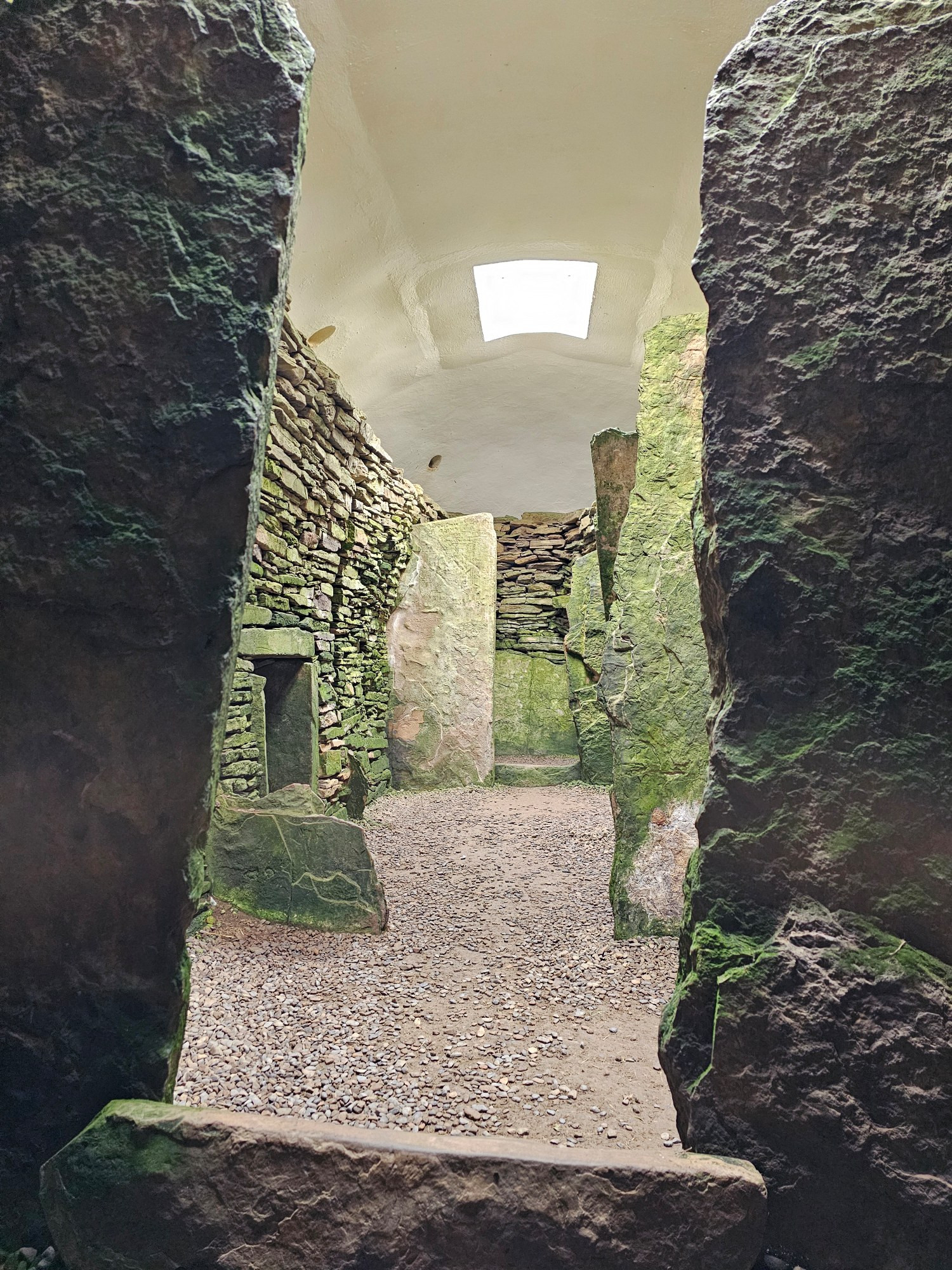 The inside of a Neolithic tomb, with tall upright stones set into the walls on either side, with a low opening on the left, and a modern roof at the top