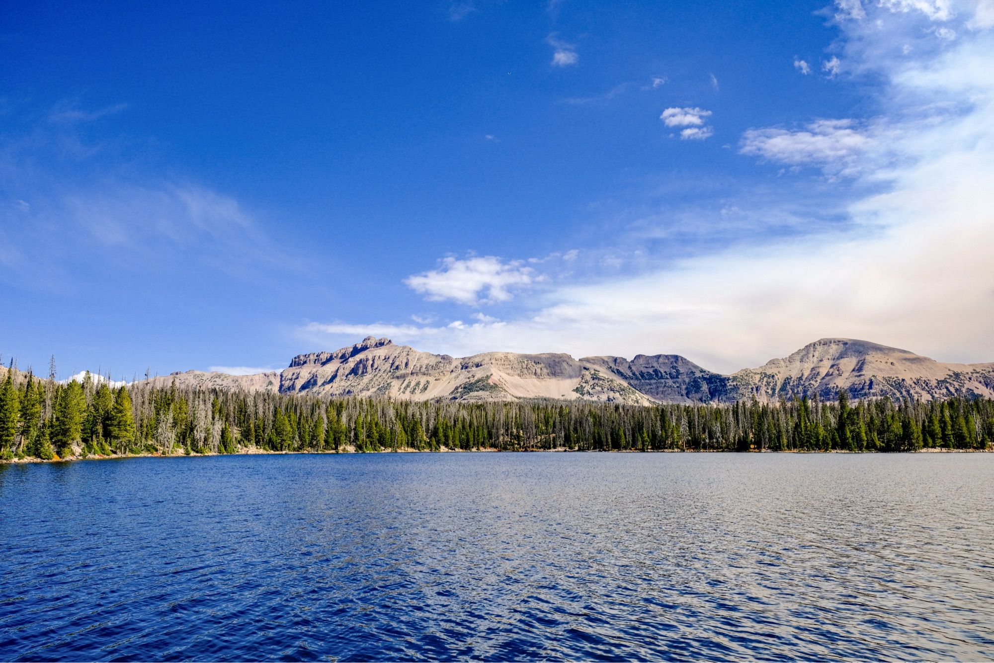 A dark blue lake with green pine trees on the shore, and bare gray rocky mountains behind the trees. The sky is blue with some white clouds and a little smoke