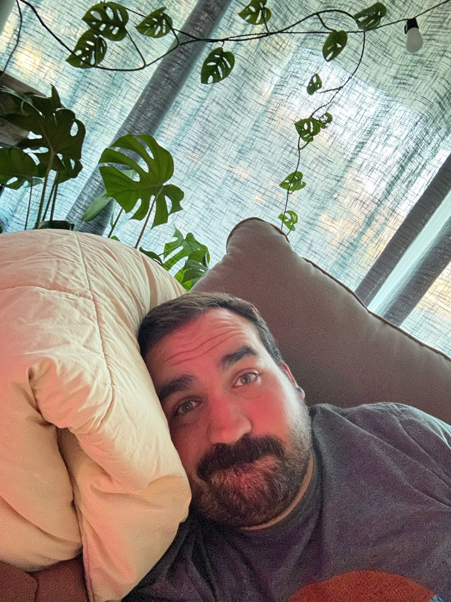 A white man with a mustache and short beard lying down on a couch staring blankly at the camera. Two monstera plants are in the background