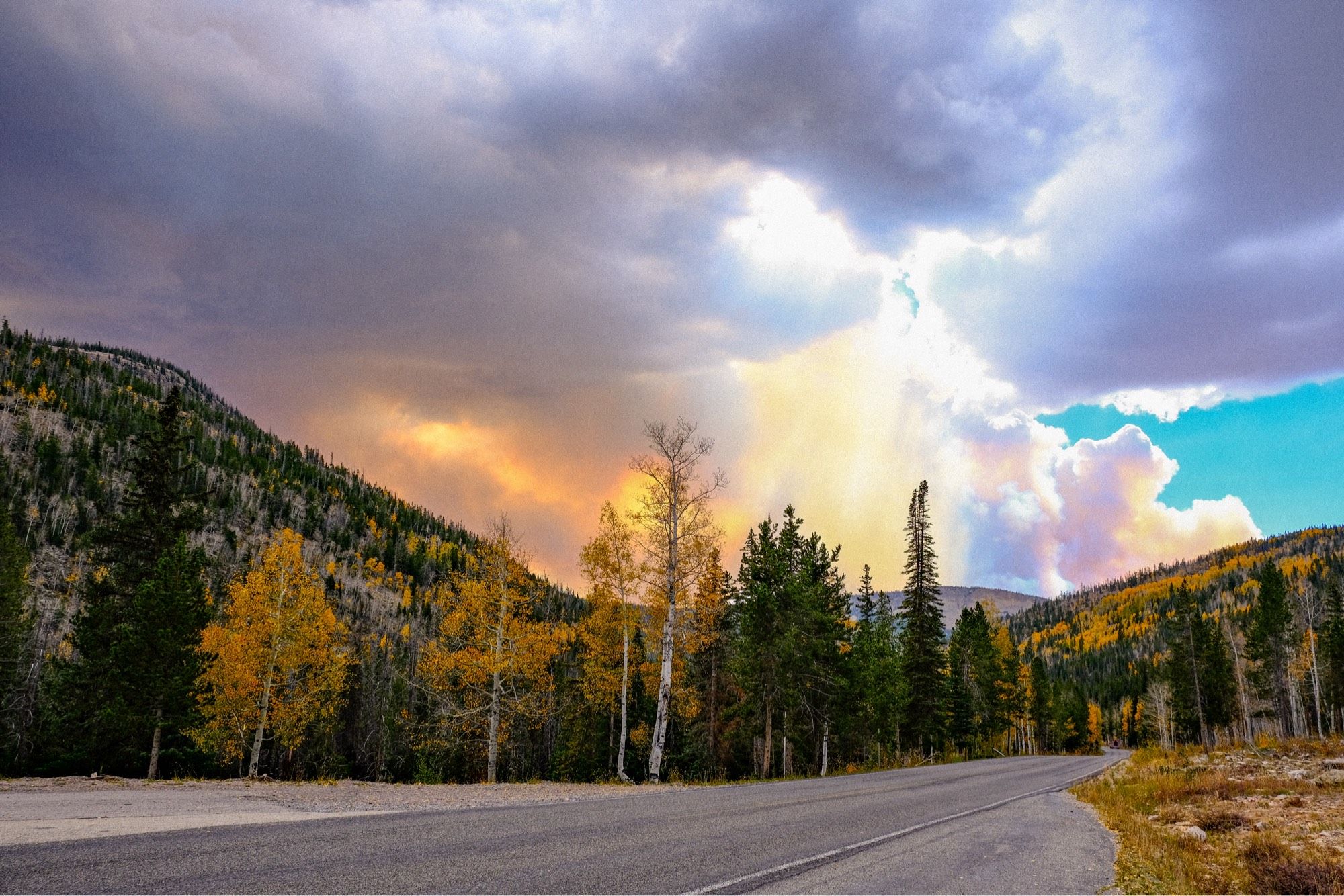 Mountains with green pine trees and yellow aspens on either side of a road. Dark clouds and orange smoke mix in the sky. A small peek of blue sky is in the middle right hand side of the picture