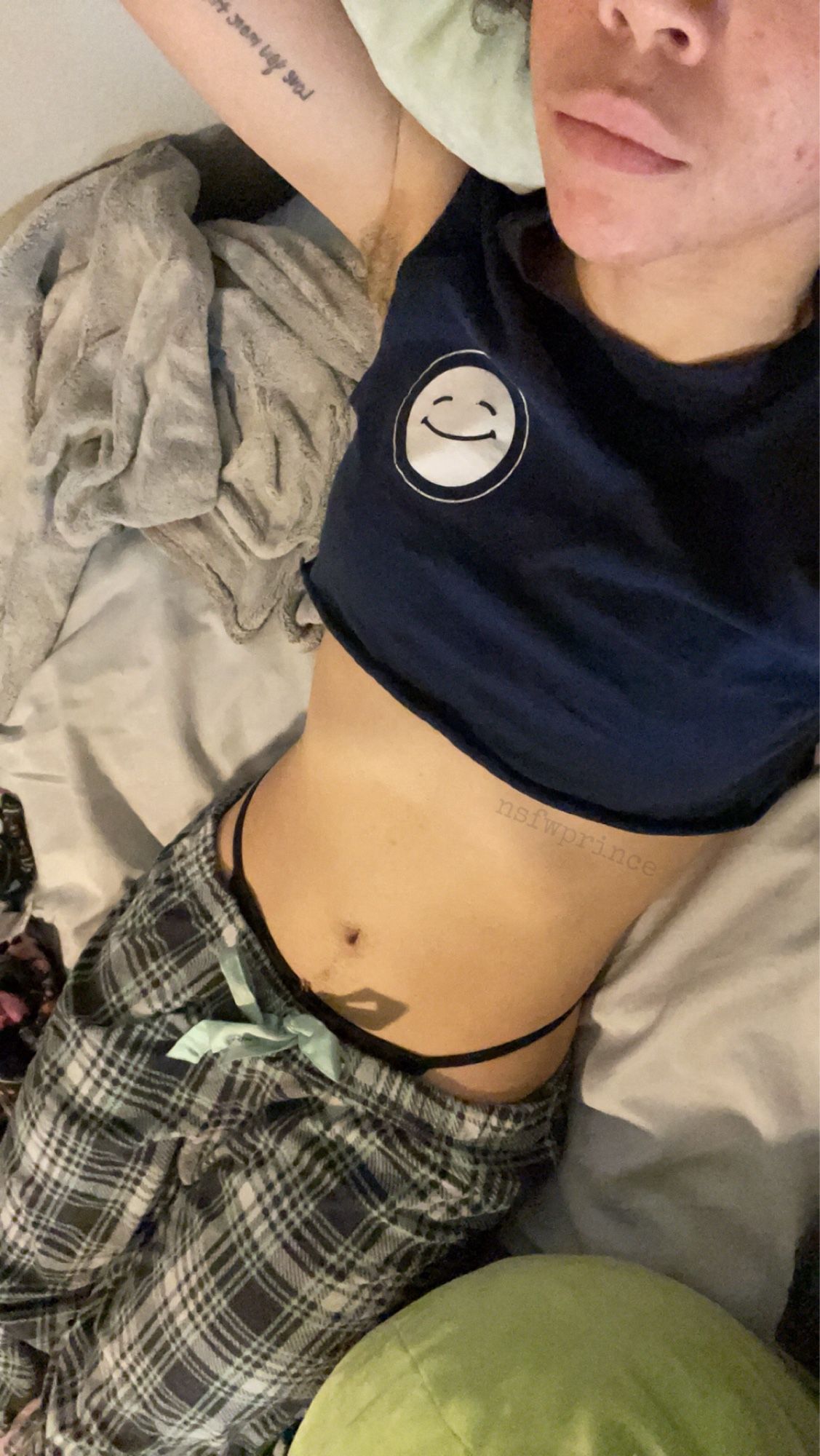 angel laying in bed in green plaid pajama pants and a navy blue crop top with a smiley face on it. they are showing off their cute tummy and black g string with their pouty lips on display