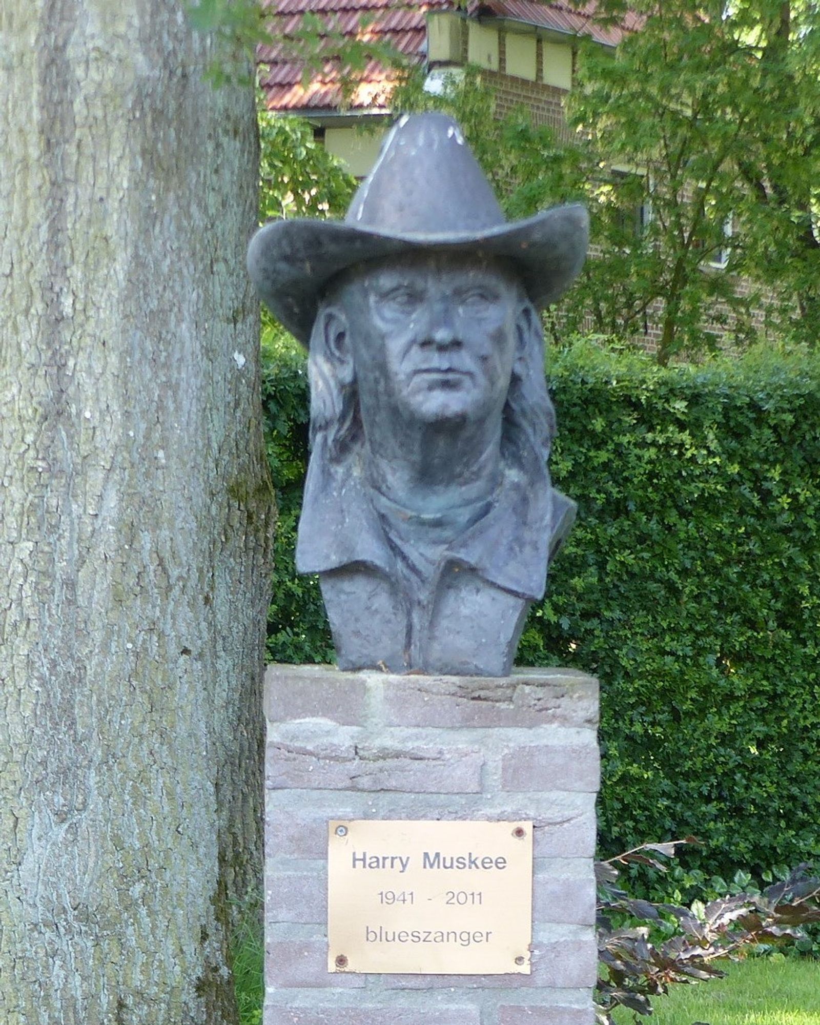 Beeld van een mannenfiguur met cowboyhoed op. De man heeft lang haar. Op de gemetselde sokkel een bord met de tekst: Harry Muskee, 1941-2011, blueszanger.
Links van het beeld een dikke boomstam, erachter een groene heg.