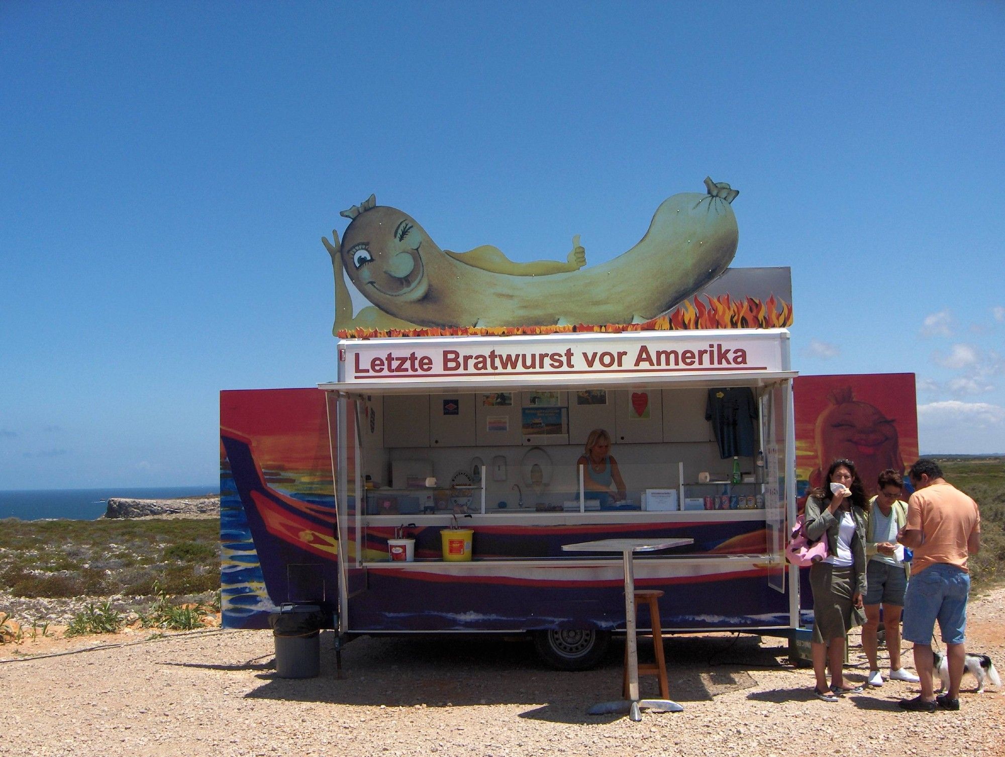 Eetkar in Cabo de Sao Vicente, Portugal, met op de kar een knipogende braadworst. Tekst op de kar Letzte Bratwurst vor Amerika.
Op de achtergrond rotsen met groen bedekt en de zee. Op de voorgrond etende mensen.

Food truck in Cabo de Sao Vicente Portugal, with a winking wiener on top of it, saying Letzte Bratwurst vor Amerika.
On the background cliffs and the sea. In front of the truck people eating.