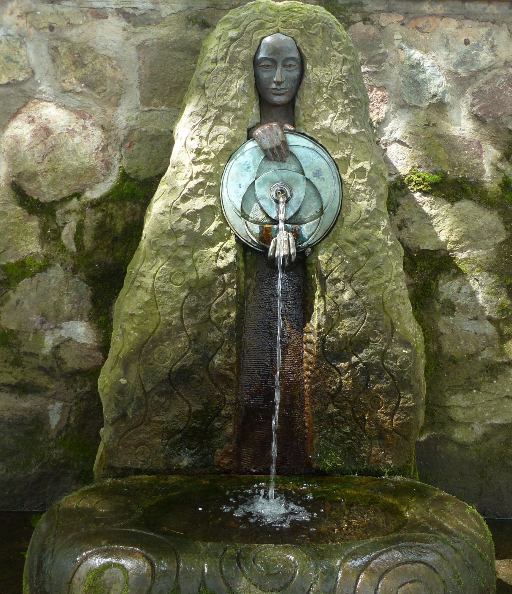 Vrouwenbeeld van steen met een soort rotsmuur erachter. Het stenen haar van de vrouw valt links en rechts tot op de grond. Hier is een versierde stenen wateropvangbak. De vrouw houdt een rond schild voor zich waaruit water stroomt.