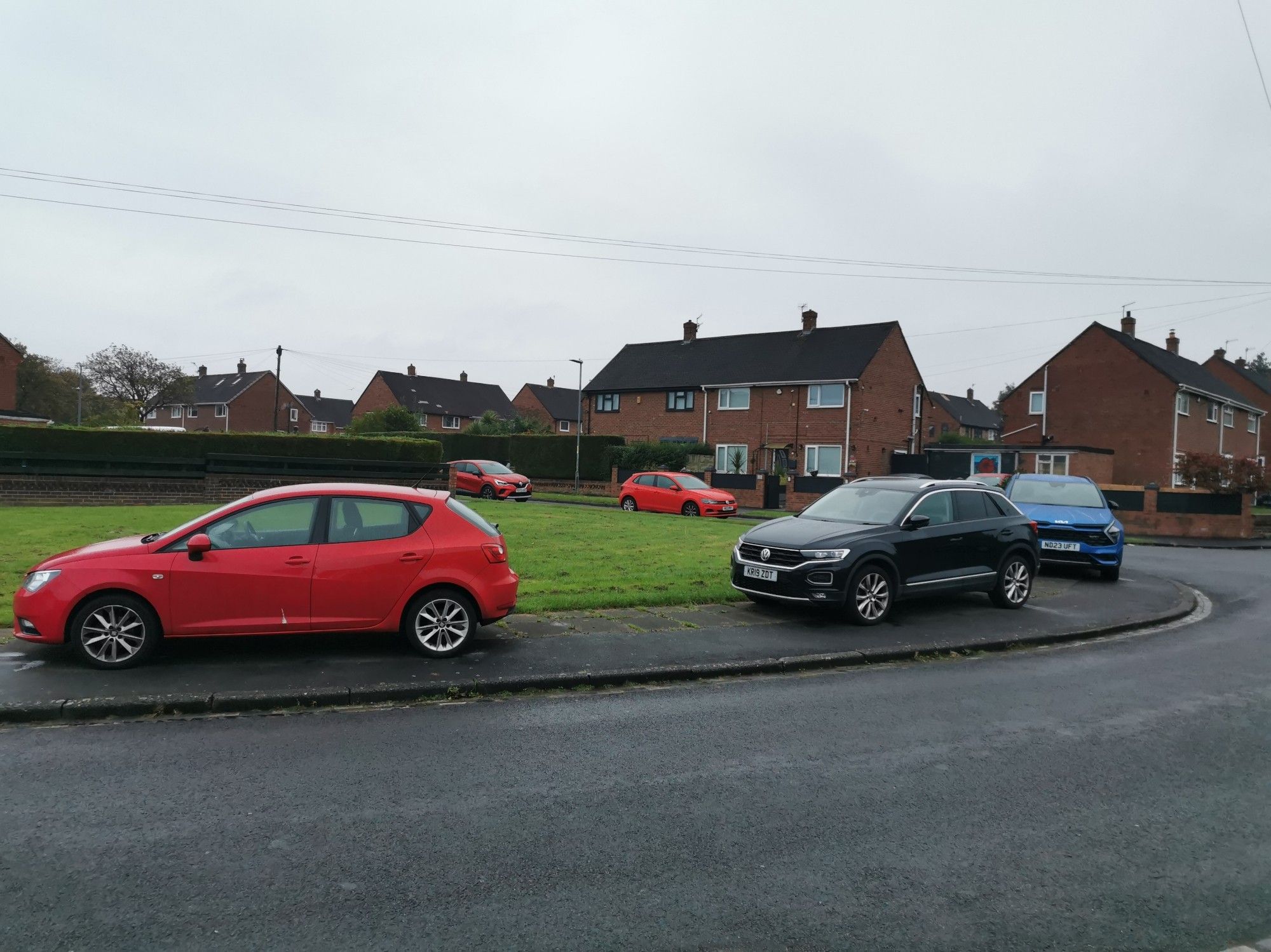 A red car, a black cat and a blue car all parked entirely on the pavement. Most people could walk past (very close to the road but without leaving the pavement) but it is impassible with most wheelchairs, pushchairs, etc. Corner on a residential street.