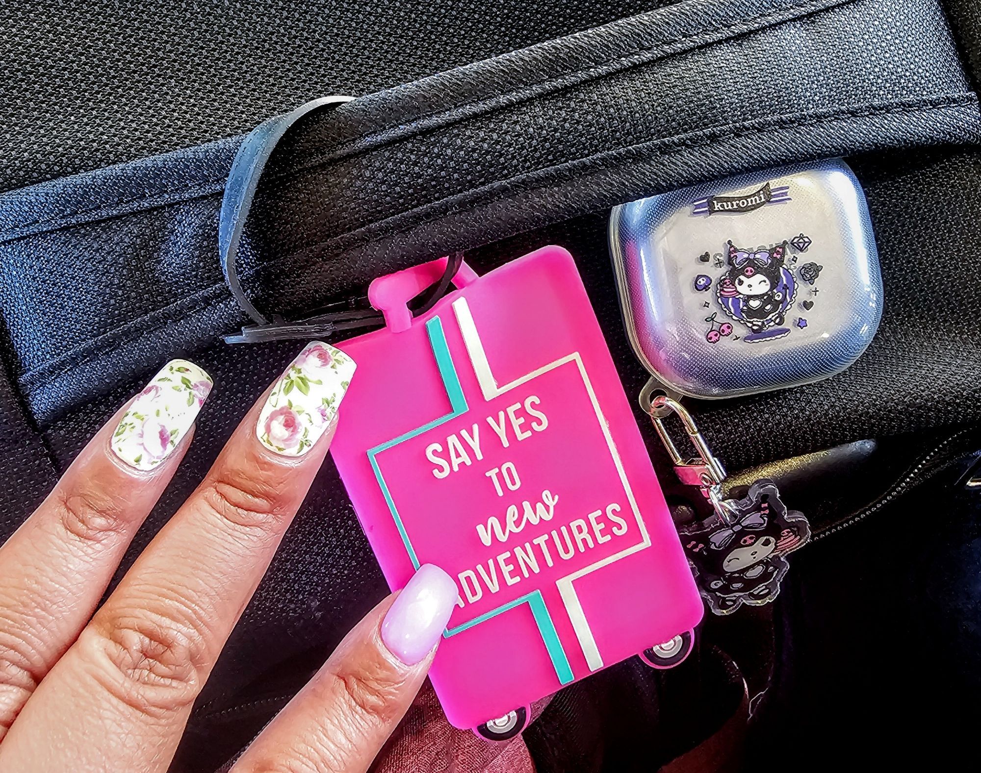 My nails with a floral design and one nail with pink. A suitcase tag that says say yes to adventures. On the suitcase is my kuromi earbud case.