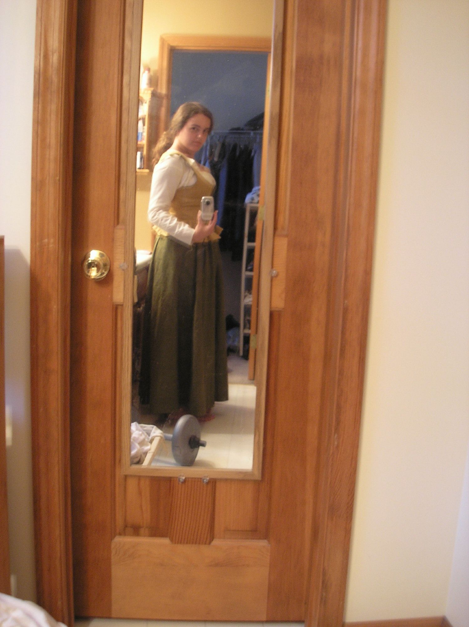 The reflection of a young brunette in a green petticoat and gold-colored bodice, holding a camera to take a shaky selfie in a mirror.