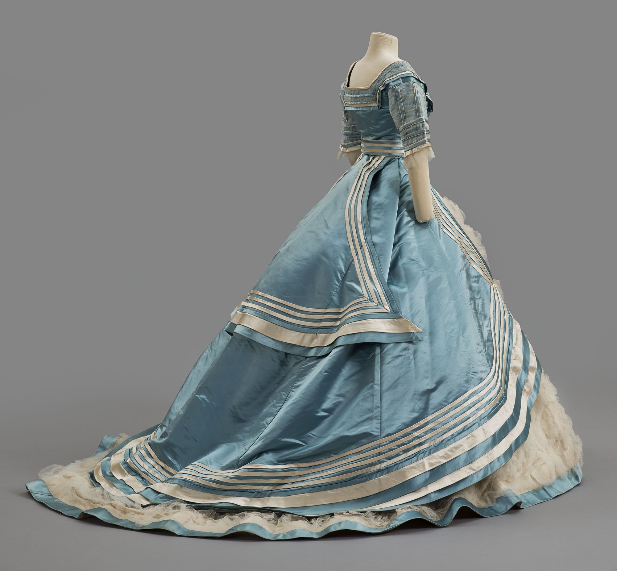 A ca. 1867 evening dress of pale blue silk satin, trimmed with stripes of white satin cut on the bias. The front of the skirt is covered with rows of ruched white tulle.