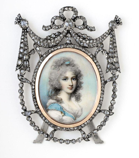 A miniature portrait of a young woman with powdered hair, wearing a white gown with a blue ribbon and a strand of pearls draped across it. There is a comb in her hair that matches the ribbon. It is set into a frame of diamonds , with a kind of bow at the top.