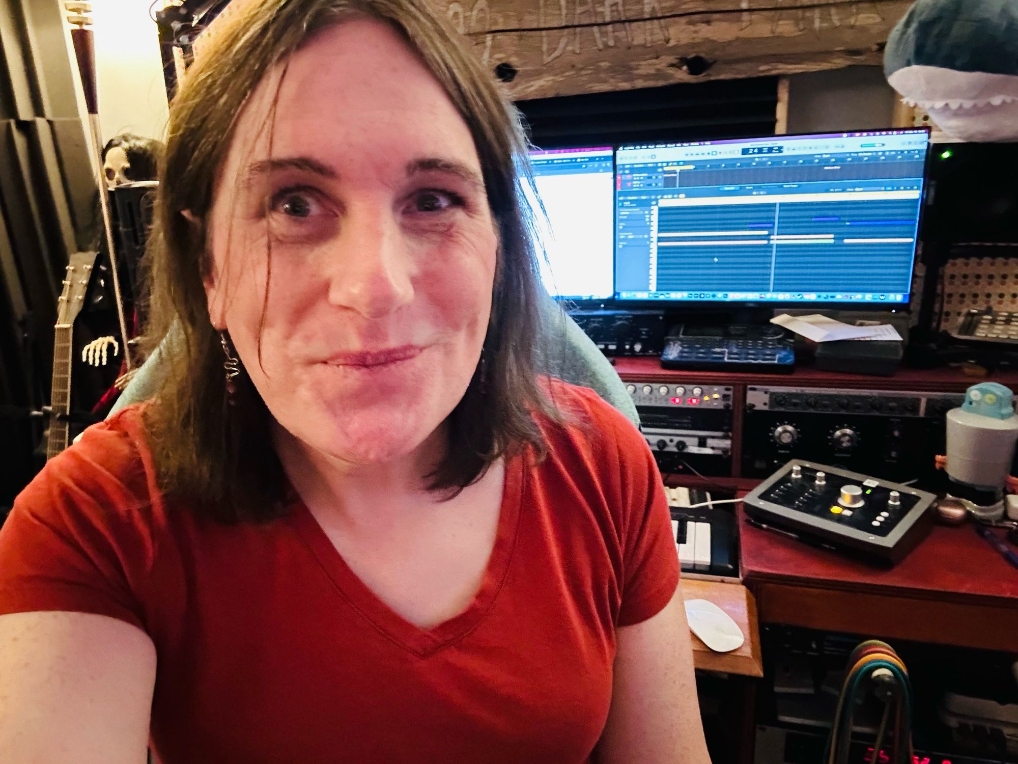 This is a selfie of Emily and her Recording studio. Emily is a white trans woman in her mid 40s here she looks a little tired is a very silly face behind her. You can see a recording, studio desk and equipment. This is logic pro running on AUDIENT hardware using a Mac mini M2 Pro. You can also see her shark Elise and a couple of skeletons around.