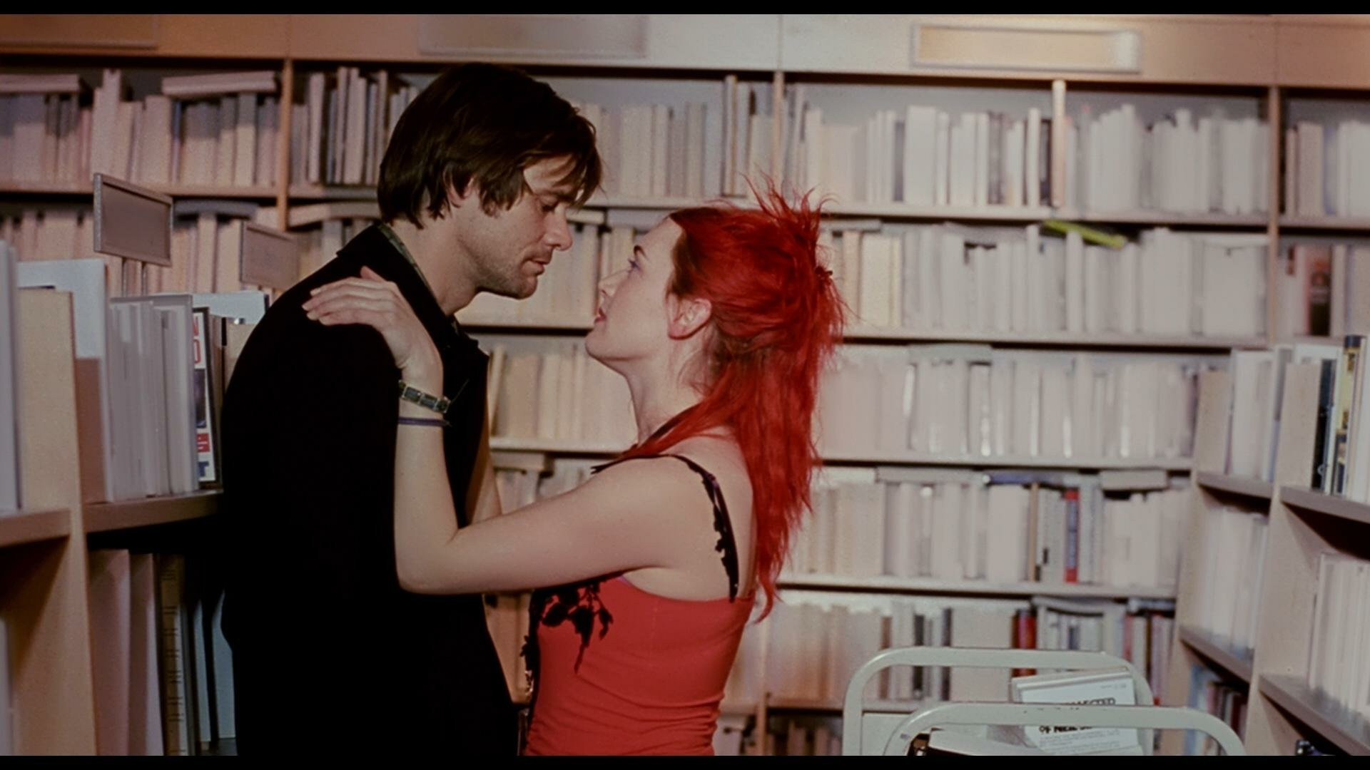 A still from Eternal Sunshine of the Spotless Mind. In it, Jim Carey and Kate Winslet embrace in a bookstore where all the covers of the books are going blank. She has bright red hair and a red shirt, he wears a black coat or shirt with long sleeves.