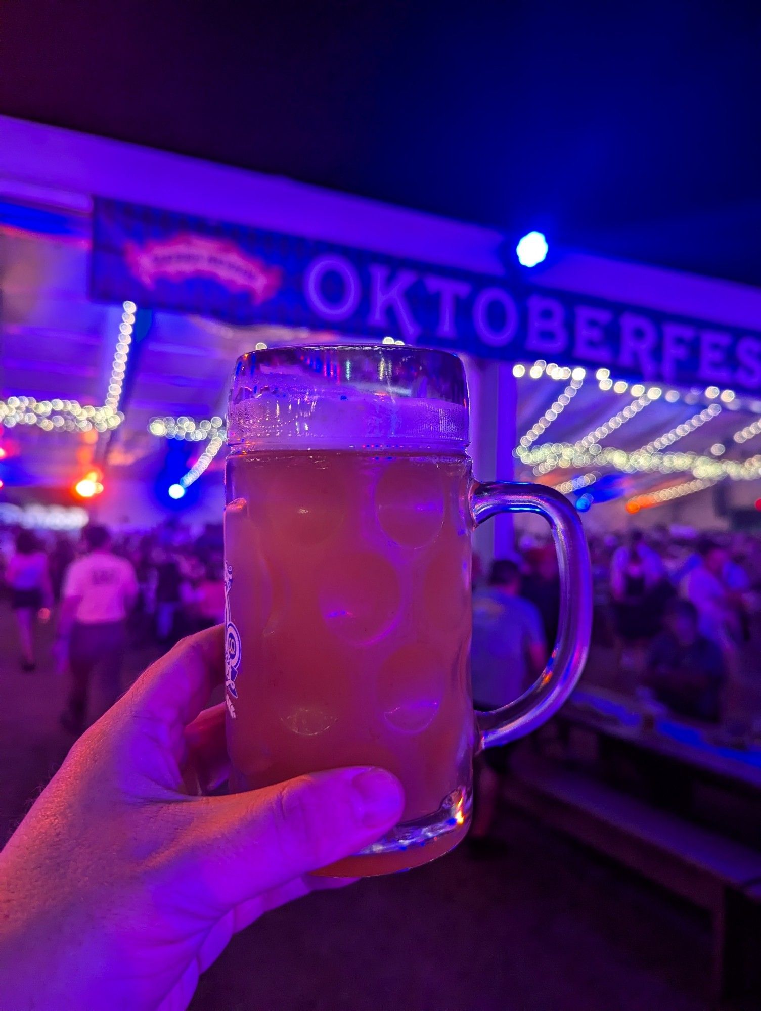 A picture of Michael J. Riser holding up a glass pint of beer in front of a busy, purple-and-red lit hall with an Oktoberfest sign hanging above it.