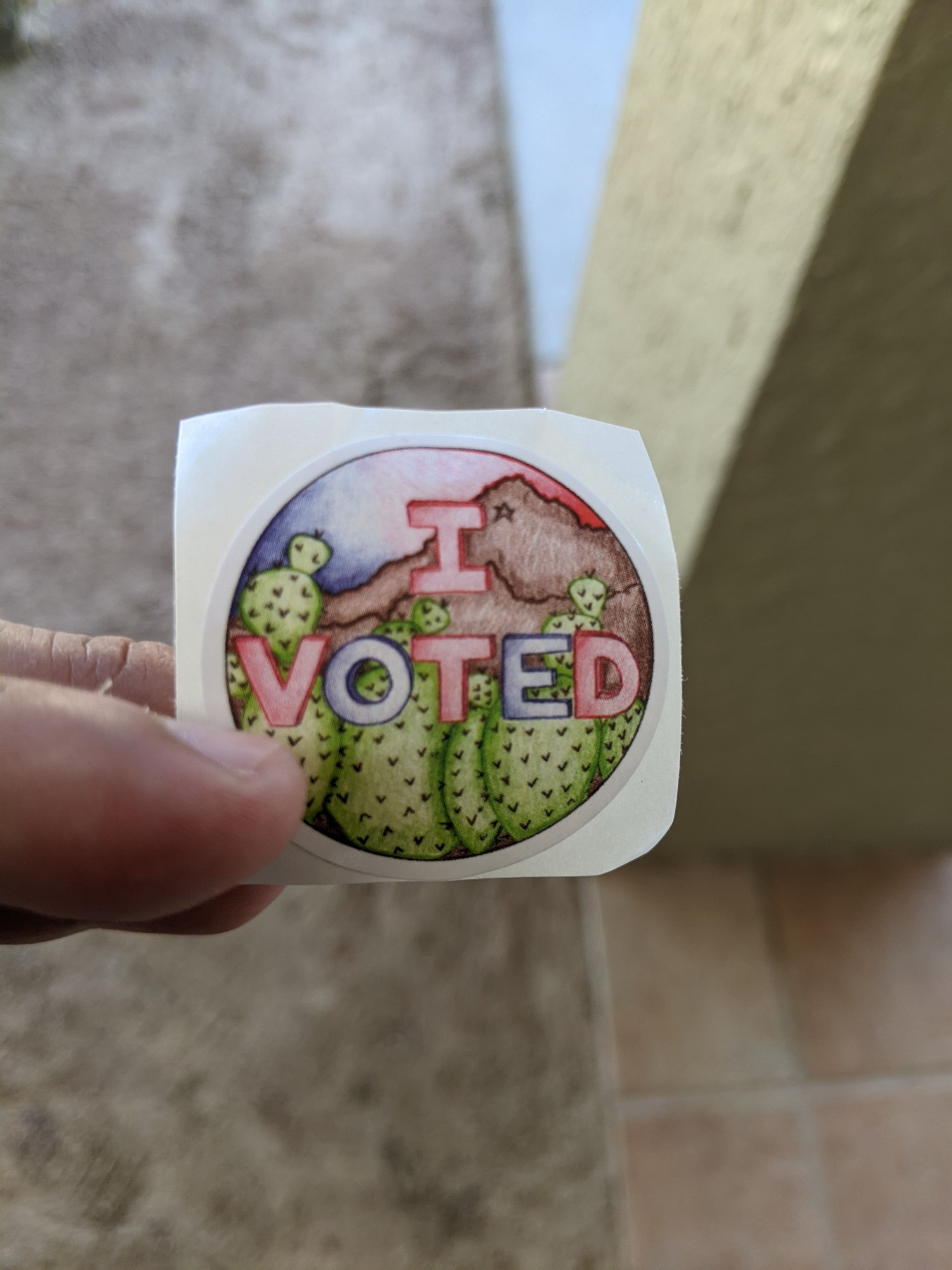 A small "I VOTED" sticker being held in the fingers of a man's hand.

The sticker features a drawing of nopales with a background of the Franklin Mountains, underneath a red, white, and blue sky.

The words "I voted" are also colored in red and blue.