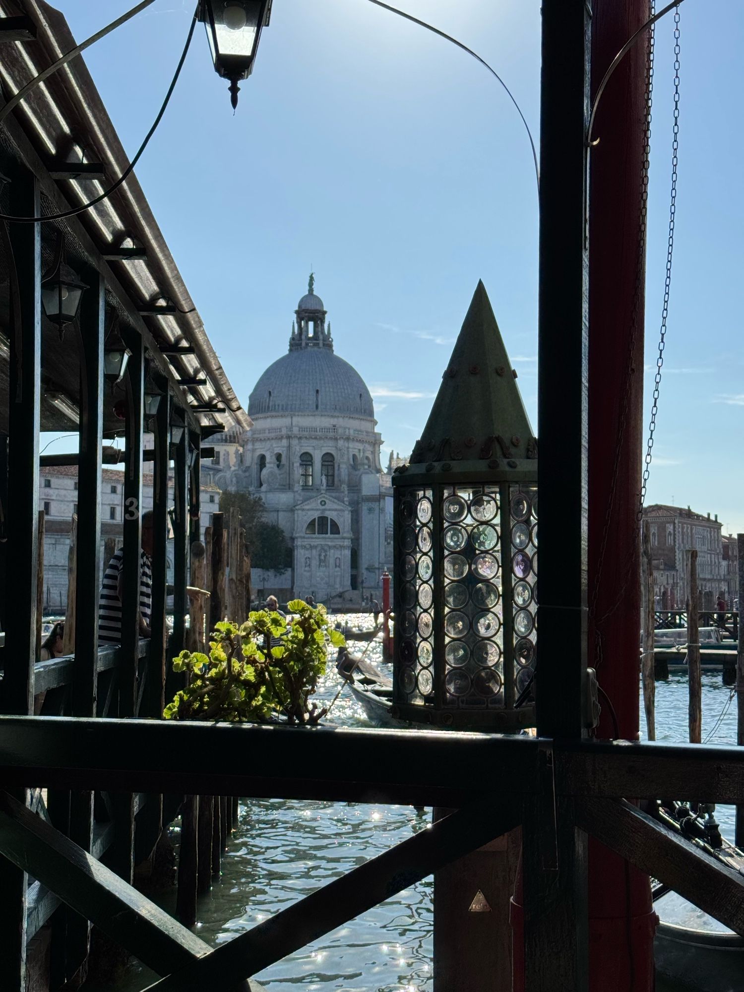 A photo taken in Venice. The picture shows the wooden fence of a pier we were waiting at to cross the main canal. Sunlight is reflecting in the water and in an unlit candle box made out of glass. In the background the Santa Maria chapel is depicted