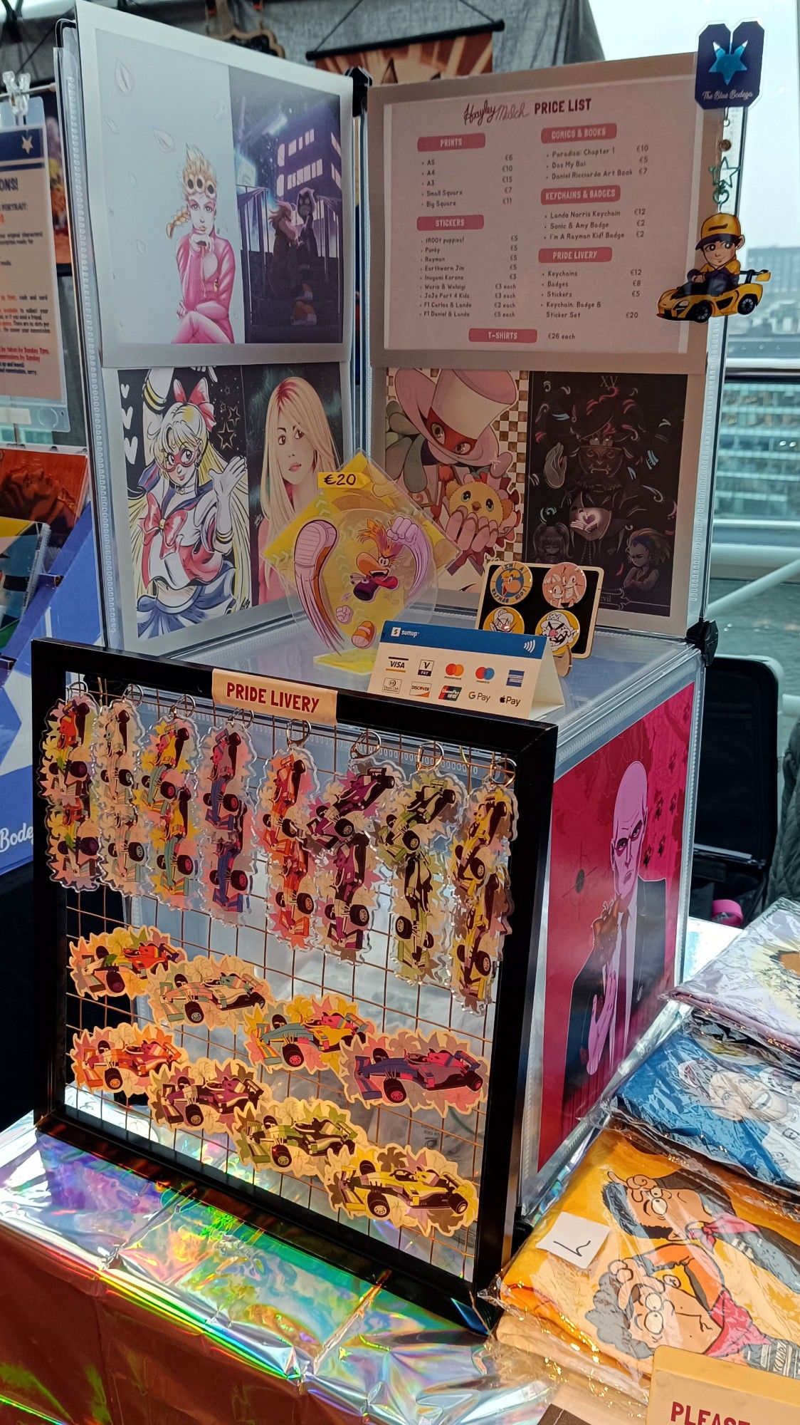 Photo of the left side of my table at the DCC trade hall, featuring prints, stickers, badges, a standee and keychains.