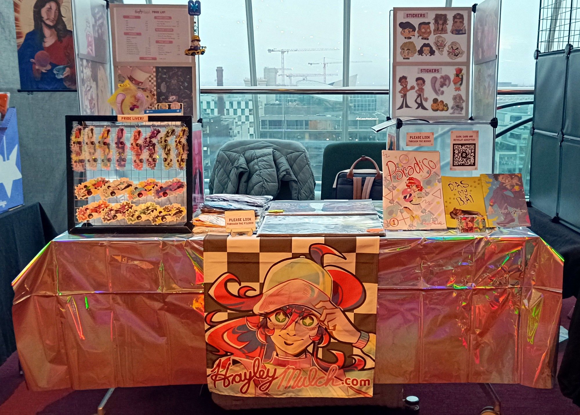 Photo of my full table at the DCC trade hall, featuring prints, stickers, badges, keychains, comics, a standee and t-shirts.