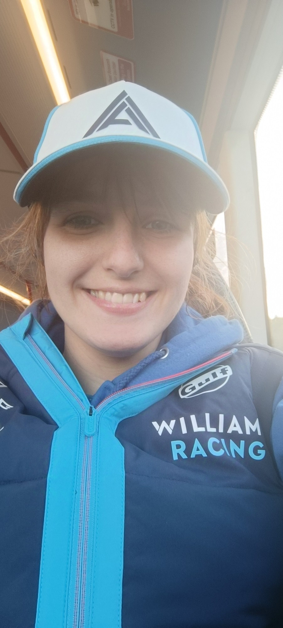 Selfie of me, a white woman with brown hair in a ponytail and fringe, wearing an Alex Albon cap and Williams Racing gilet. I'm smiling towards the viewer.