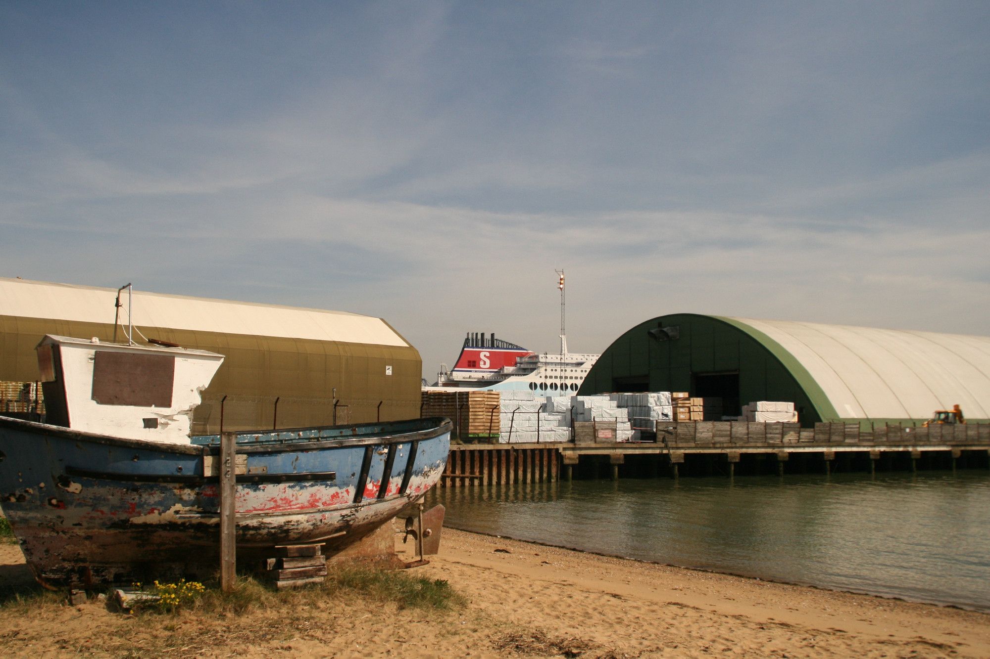 Foreground is a beach with a decaying small boat to the left. Behind that boat is a warehouse, there's another warehouse to the right behind water. A large passenger ferry passes between the warehouses.