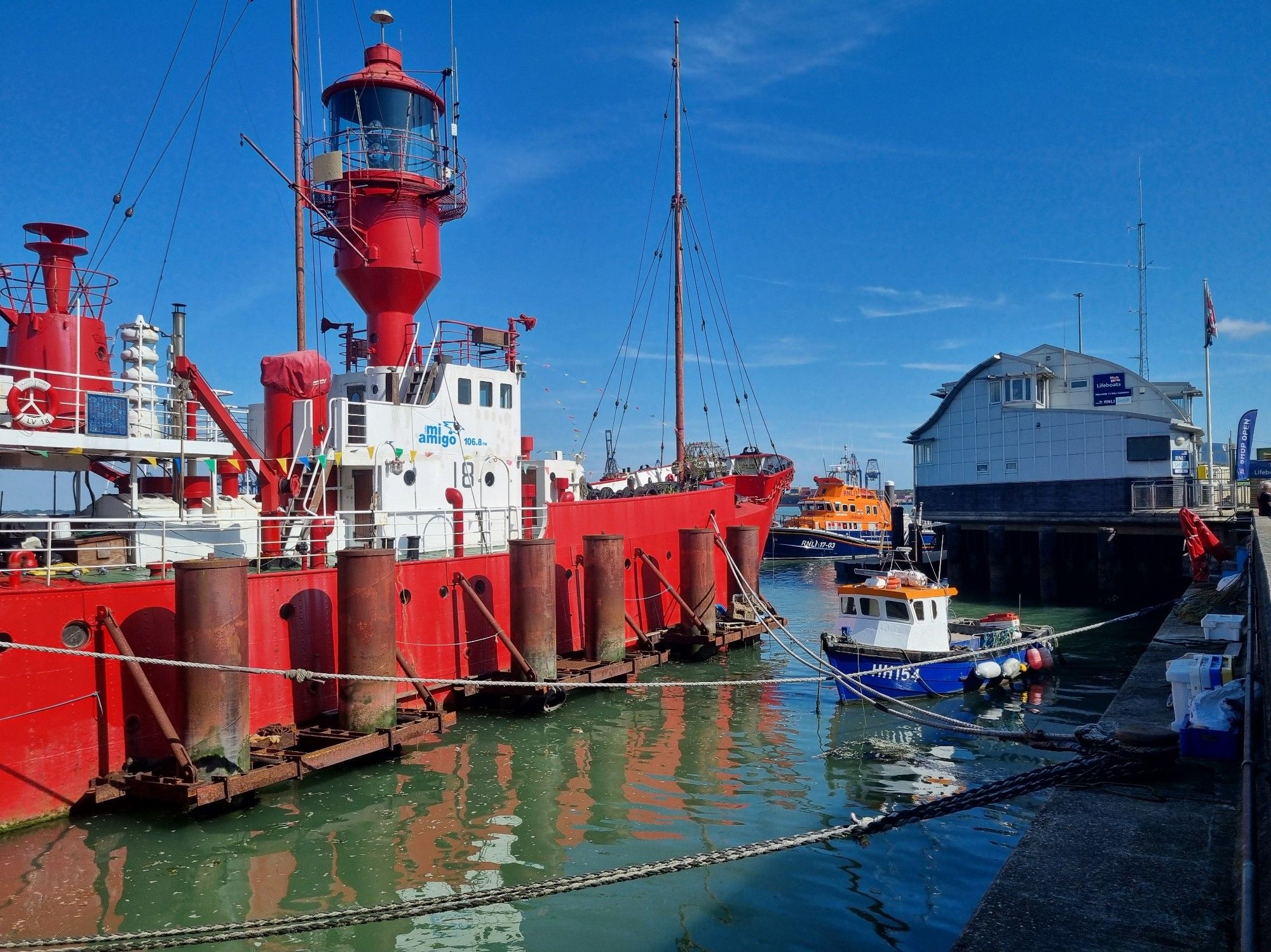 A harbour scene. A red lightship is central, behind six steel piles. A small boat is to the right, and a boat house is next to an orange lifeboat. Ropes cross the scene.