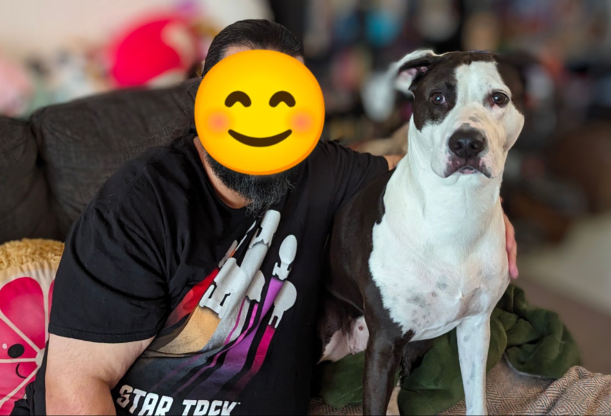 My partner wearing a Star Trek T-shirt with his arm around our dog Tasha, a black and white pitty-foxhound mix. She's standing alert and excited looking slightly to the left of the camera. He has a smiley face emoji over his face for privacy. The backgound is blurred.