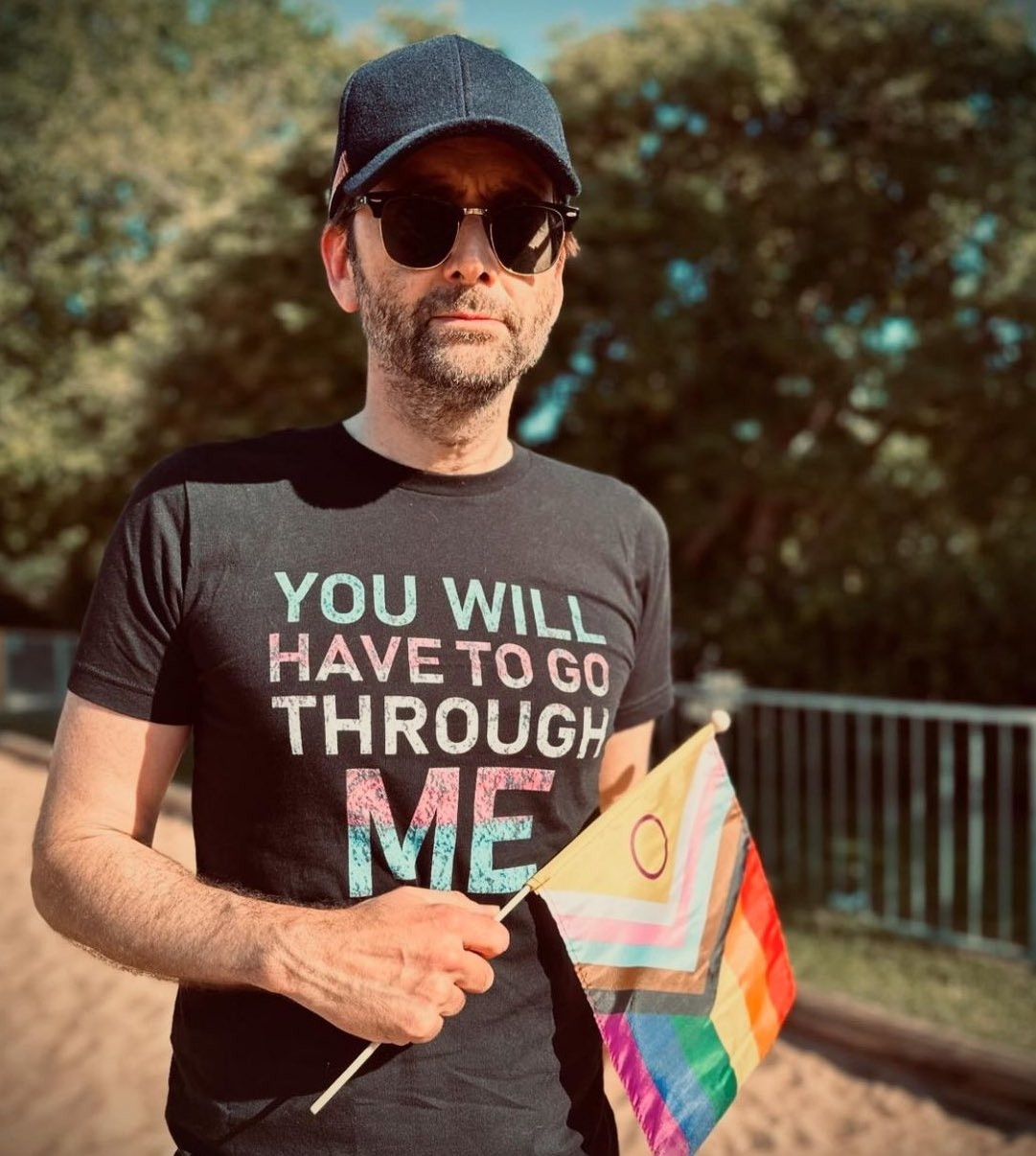 David Tennant outside wearing sunglasses, a black ball cap and a black shirt with blue, white and pink lettering (trans pride colors) that reads "YOU WILL HAVE TO GO THROUGH ME". He's also holding a small Progress Pride flag (the intersex inclusive one) up against his ribs just below the lettering of the shirt. His expression is neutral. He's slightly unshaven with a bit of salt and pepper scruff.