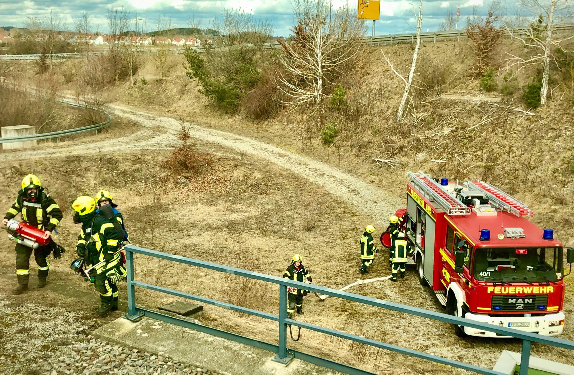 Ein Feuerwehrauto und mehrere Feuerwehrmänner in Uniform am Gleisrand, durch das Zugfenster gesehen.