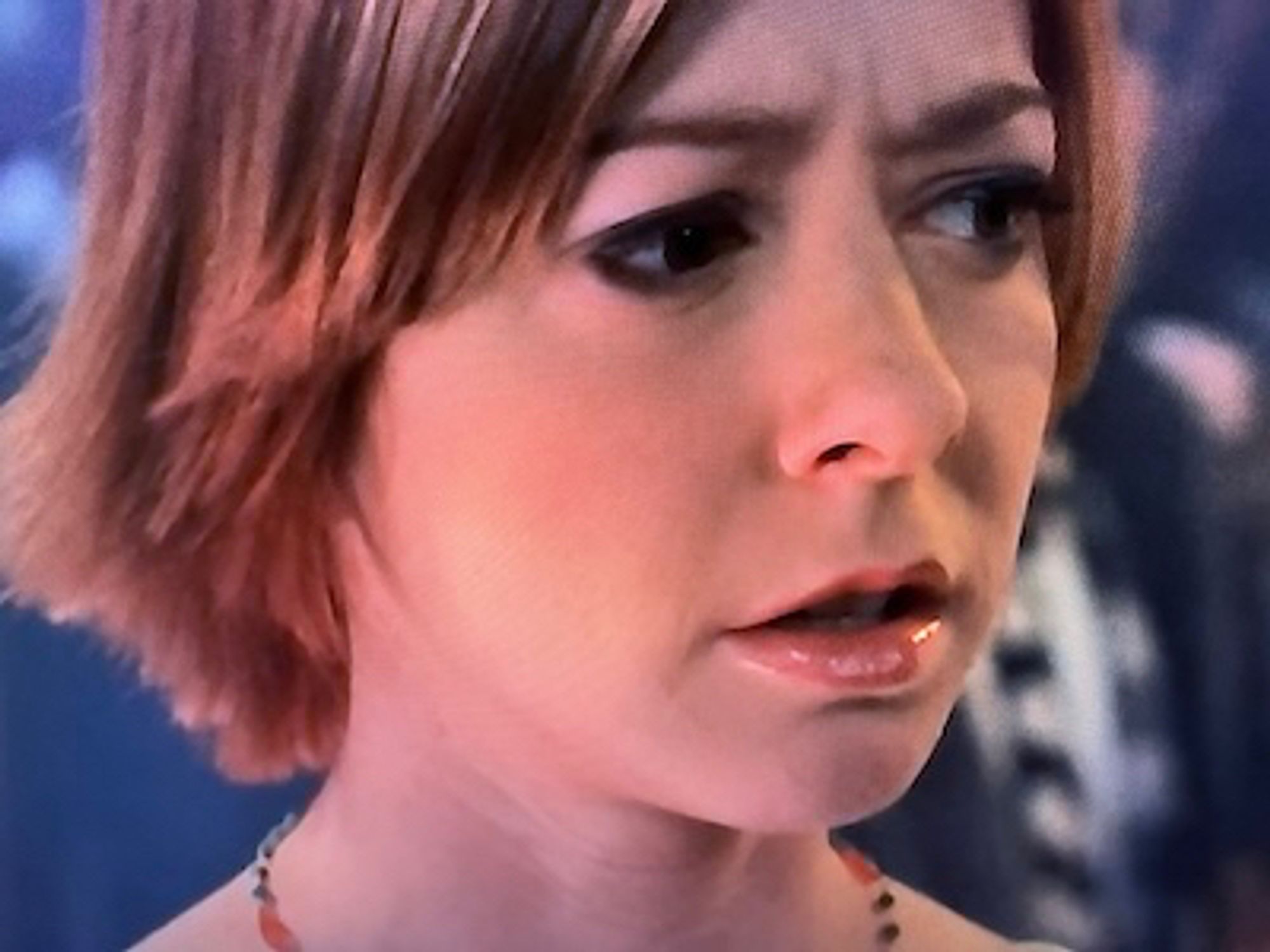 Close-up of Willow's face and neck. She has red hair, lipstick, a beaded necklace, and what seems to be eye shadow. There is someone with a black jacket and a black and white striped tie behind her on the right. Screenshot from Buffy The Vampire Slayer Season 4, Episode 22.