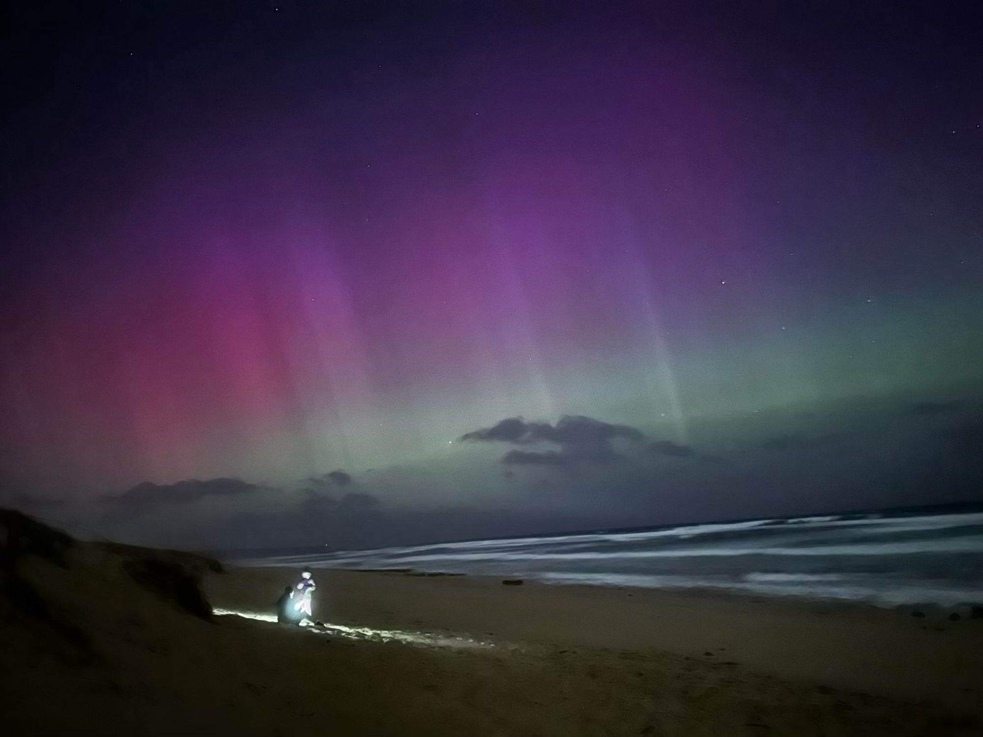 Pink and green aurora over the rollers on Port Phillip Bay. On the dark beach below, a small bright figure in brilliant torchlight is packing a bag